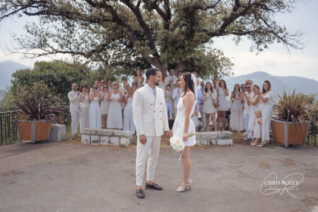 Les mariés se tiennent par la main, face à face, en plein air, à Falicon. Tous deux sont vêtus de blanc ; la mariée tient un bouquet. Derrière eux, des invités vêtus de blanc applaudissent au milieu des collines et des arbres des Alpes-Maritimes, près de Nice.