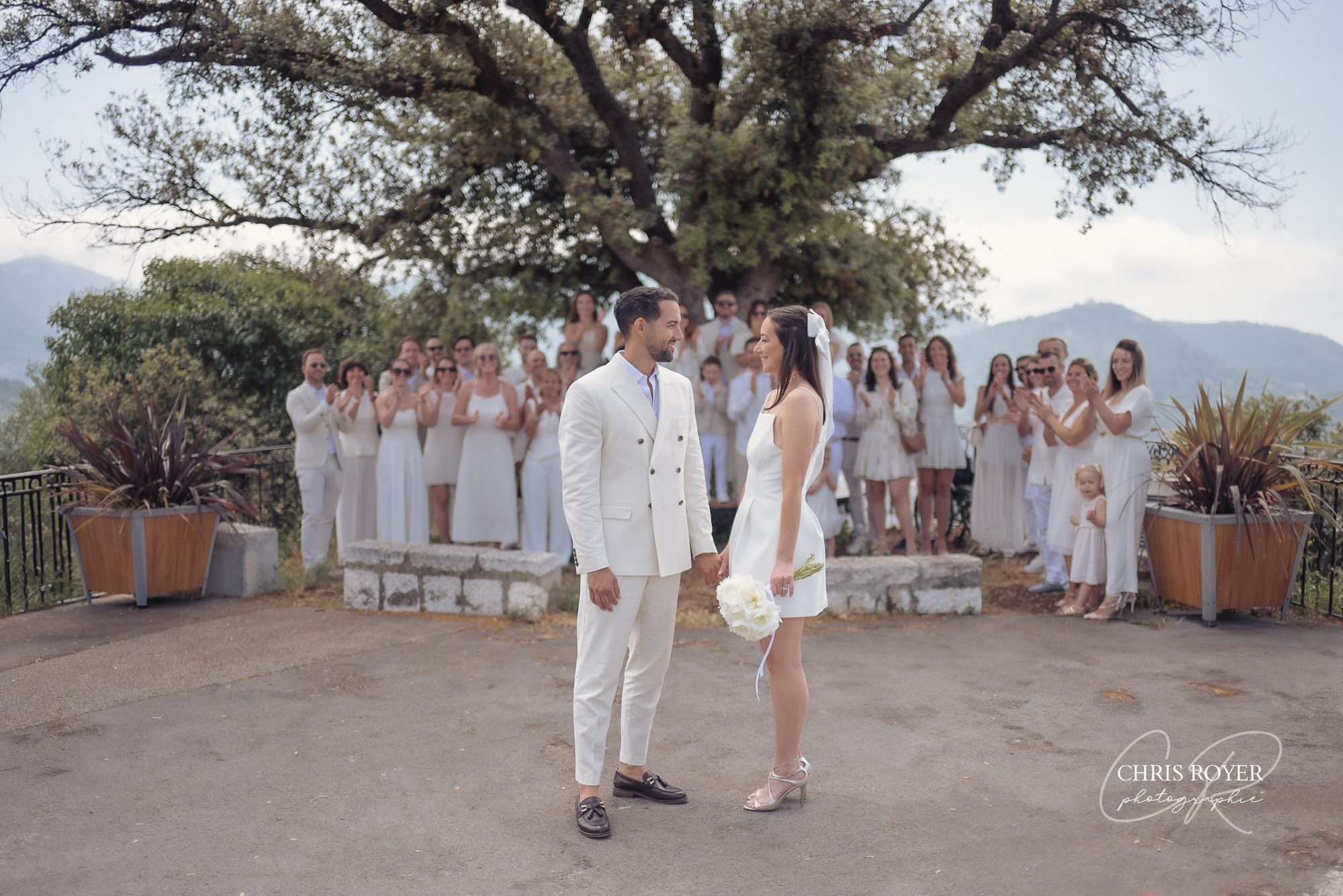 Les mariés se tiennent par la main, face à face, en plein air, à Falicon. Tous deux sont vêtus de blanc ; la mariée tient un bouquet. Derrière eux, des invités vêtus de blanc applaudissent au milieu des collines et des arbres des Alpes-Maritimes, près de Nice.