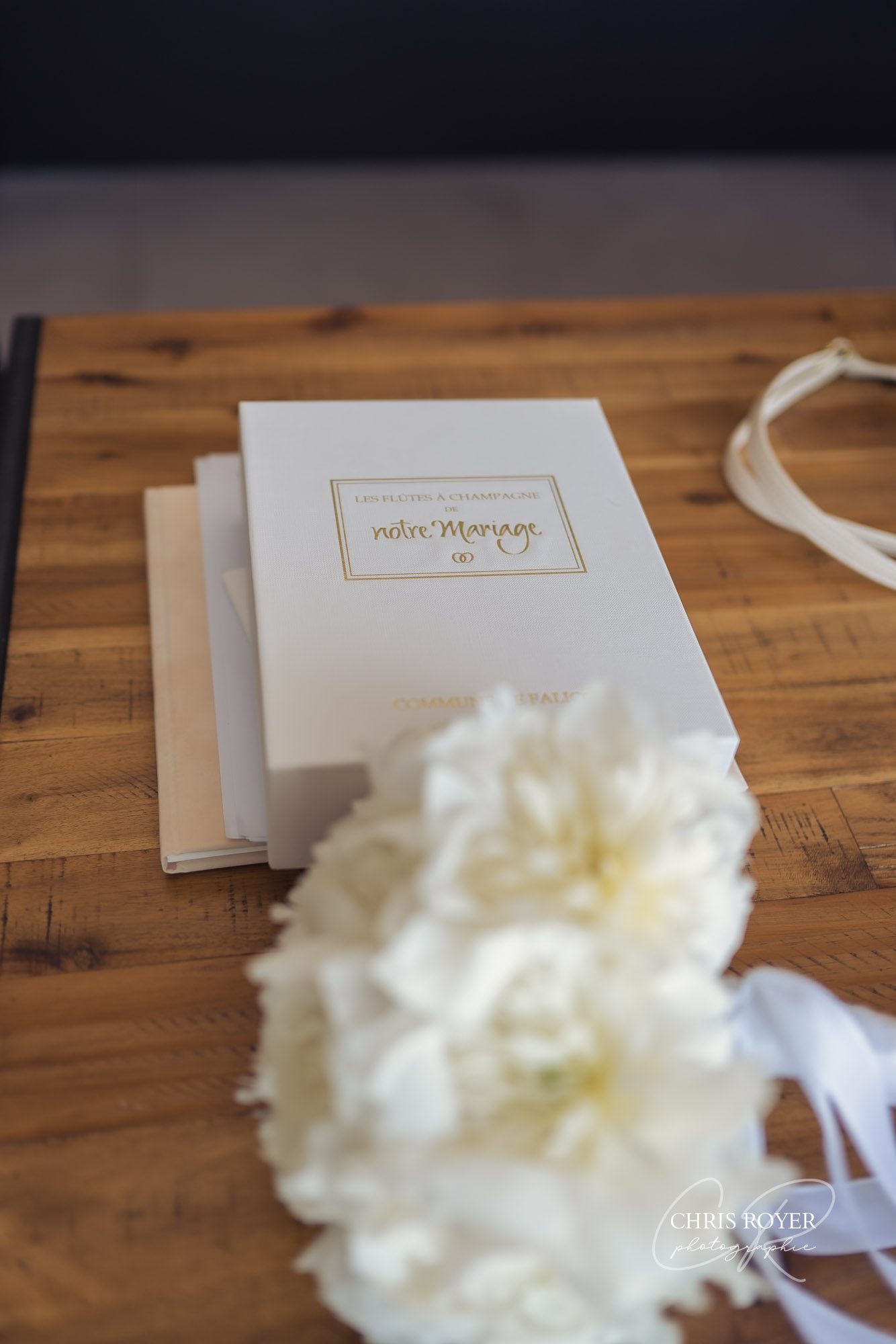 Un livre de mariage blanc étiqueté notre Mariage repose sur une table en bois à côté d'un bouquet de fleurs blanches et d'un ruban, capturant l'ambiance élégante d'un mariage à Falicon.