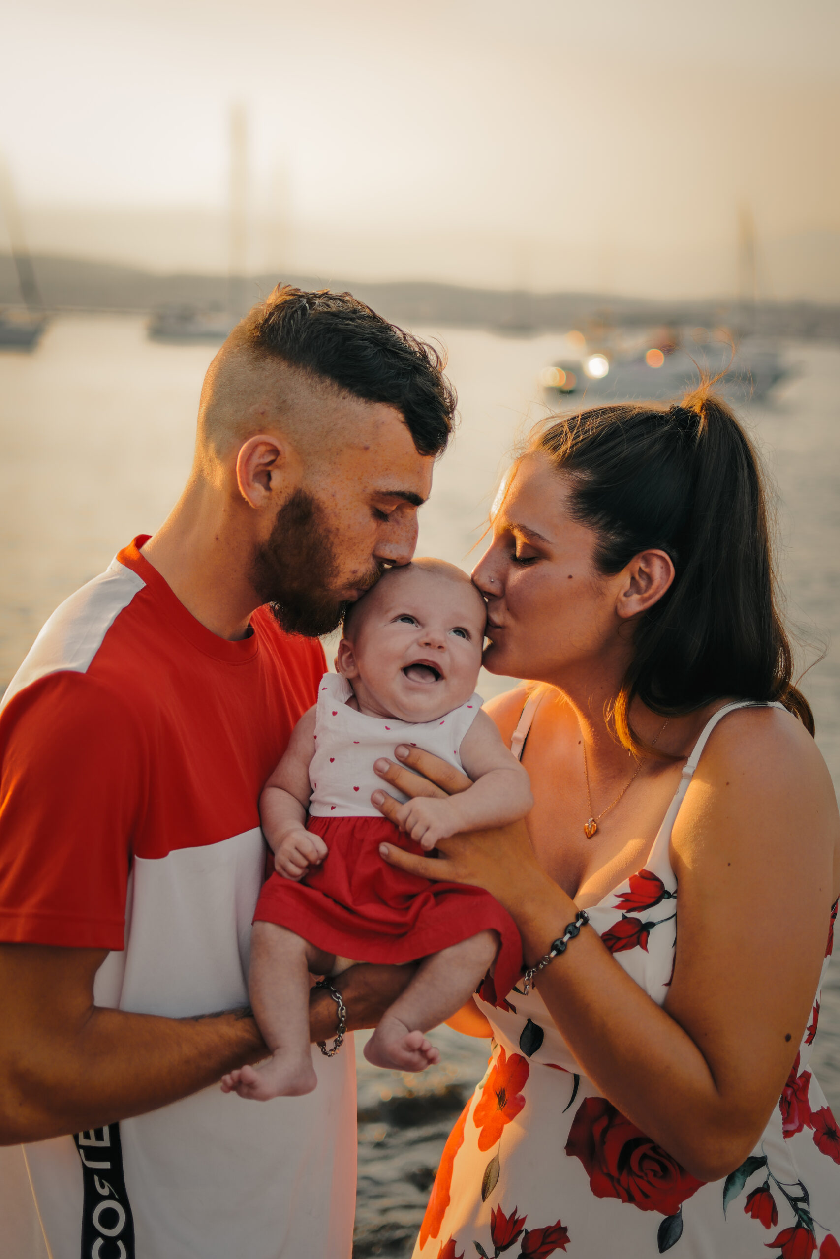 Un couple embrasse tendrement leur bébé souriant alors qu'il se tient en plein air près de la mer au coucher du soleil à Antibes, avec des bateaux et un ciel brumeux en arrière-plan.