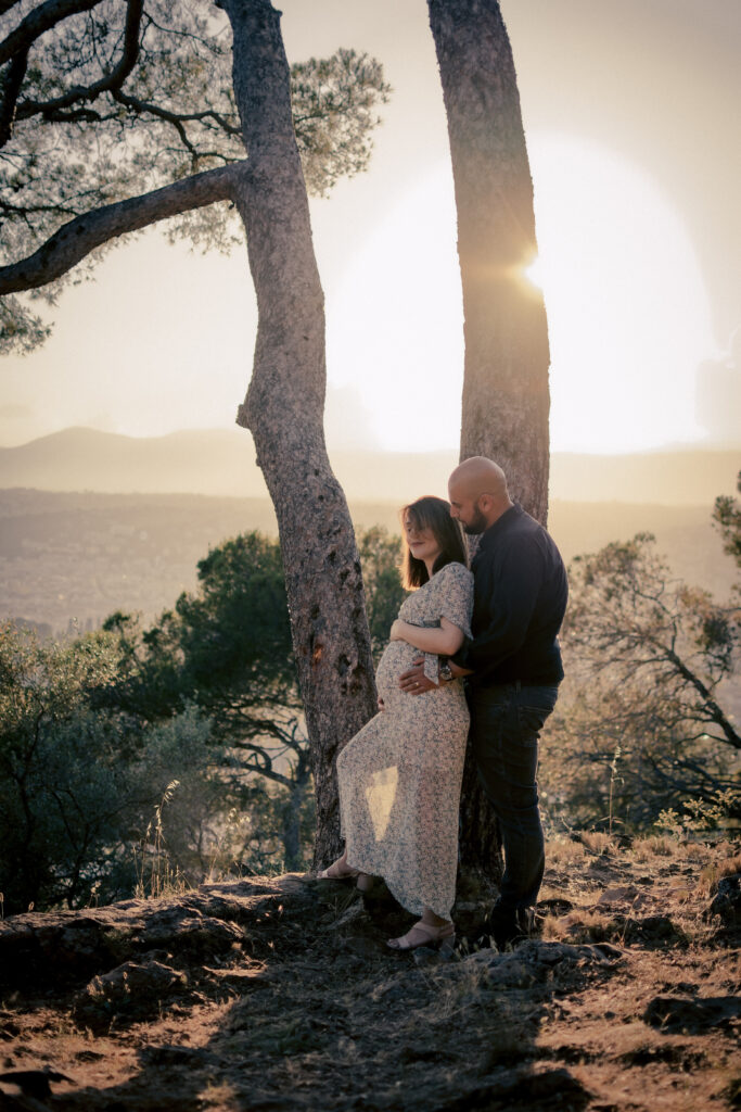 Une femme enceinte vêtue d'une robe à fleurs se tient à côté d'un homme, tous deux adossés à un arbre au coucher du soleil, avec une douce lumière dorée et les montagnes pittoresques des Alpes-Maritimes en arrière-plan, capturant la beauté de la maternité.
