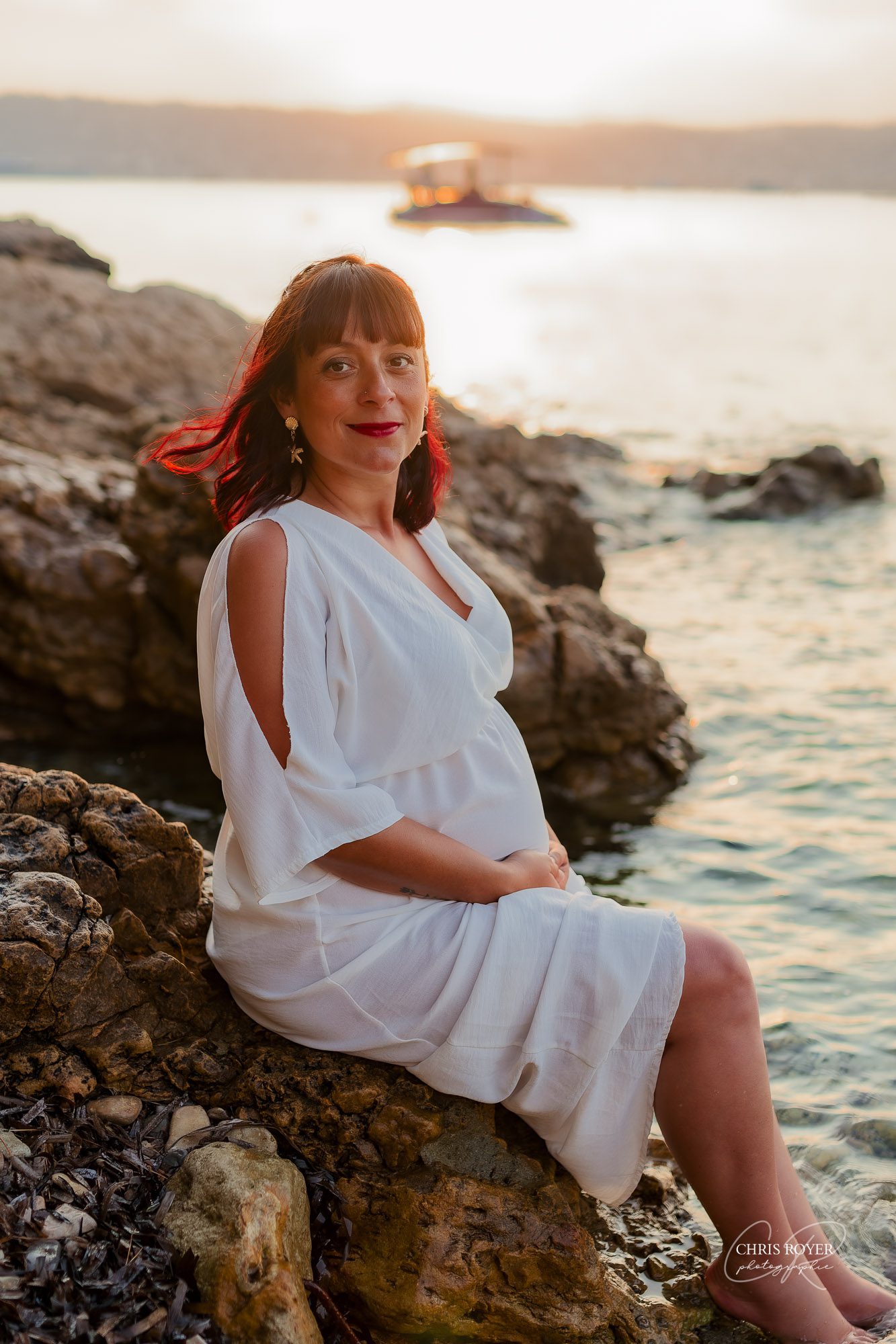 Une femme enceinte vêtue d'une robe blanche est assise sur un rivage rocheux au bord d'une eau calme au coucher du soleil, regardant l'appareil photo avec un doux sourire. Parfaite pour un portfolio sur la maternité, la scène est agréable et sereine, avec un bateau au loin et la lumière du soleil qui brille derrière elle.
