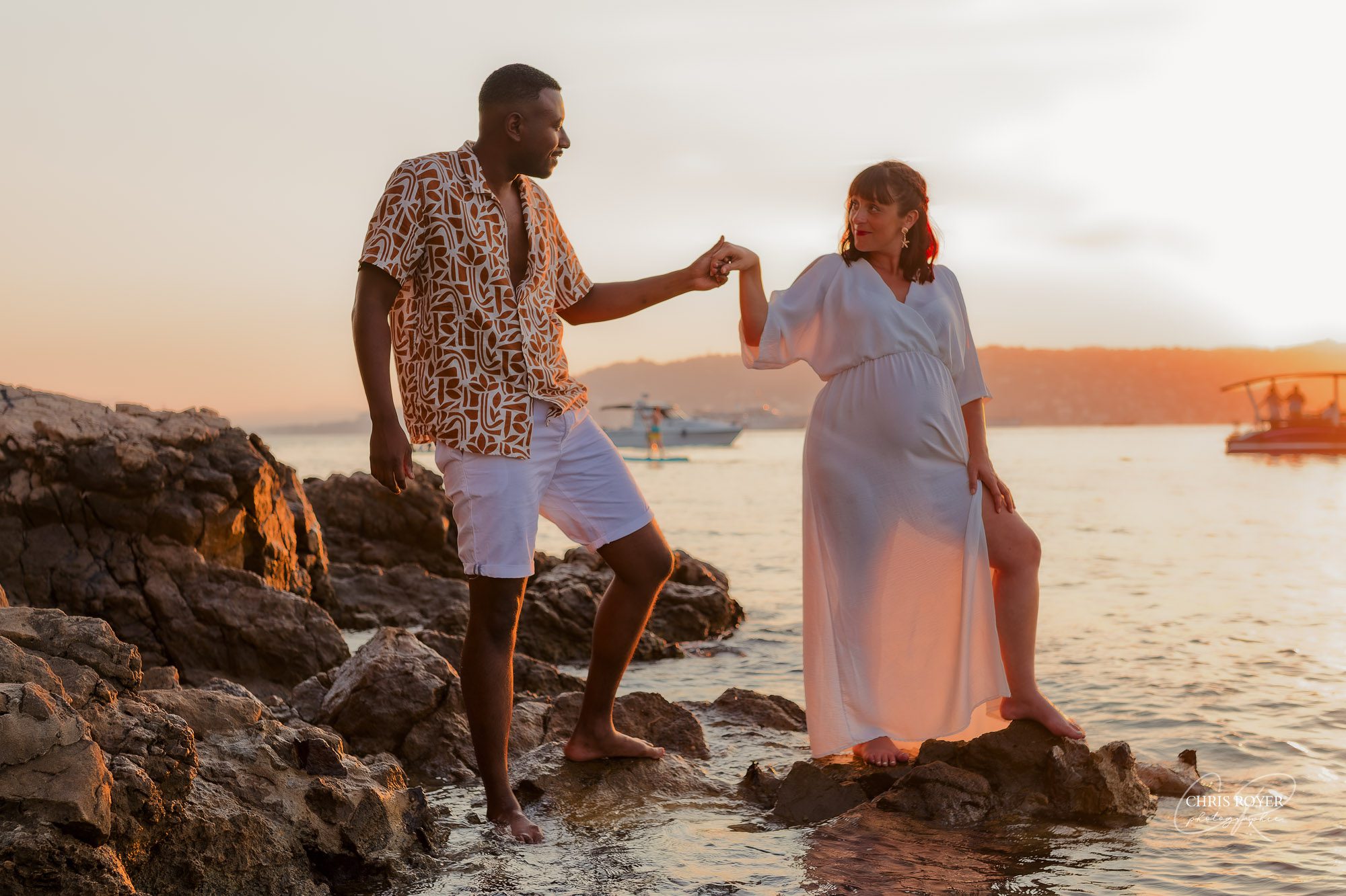 Un couple se tient sur un rivage rocheux au coucher du soleil ; l'homme, vêtu d'une chemise à motifs et d'un short, aide la femme enceinte, vêtue d'une robe blanche fluide, à se tenir en équilibre sur les rochers. Leur moment de tendresse au bord de l'eau ajoute une touche agréable à tout portfolio sur la maternité.