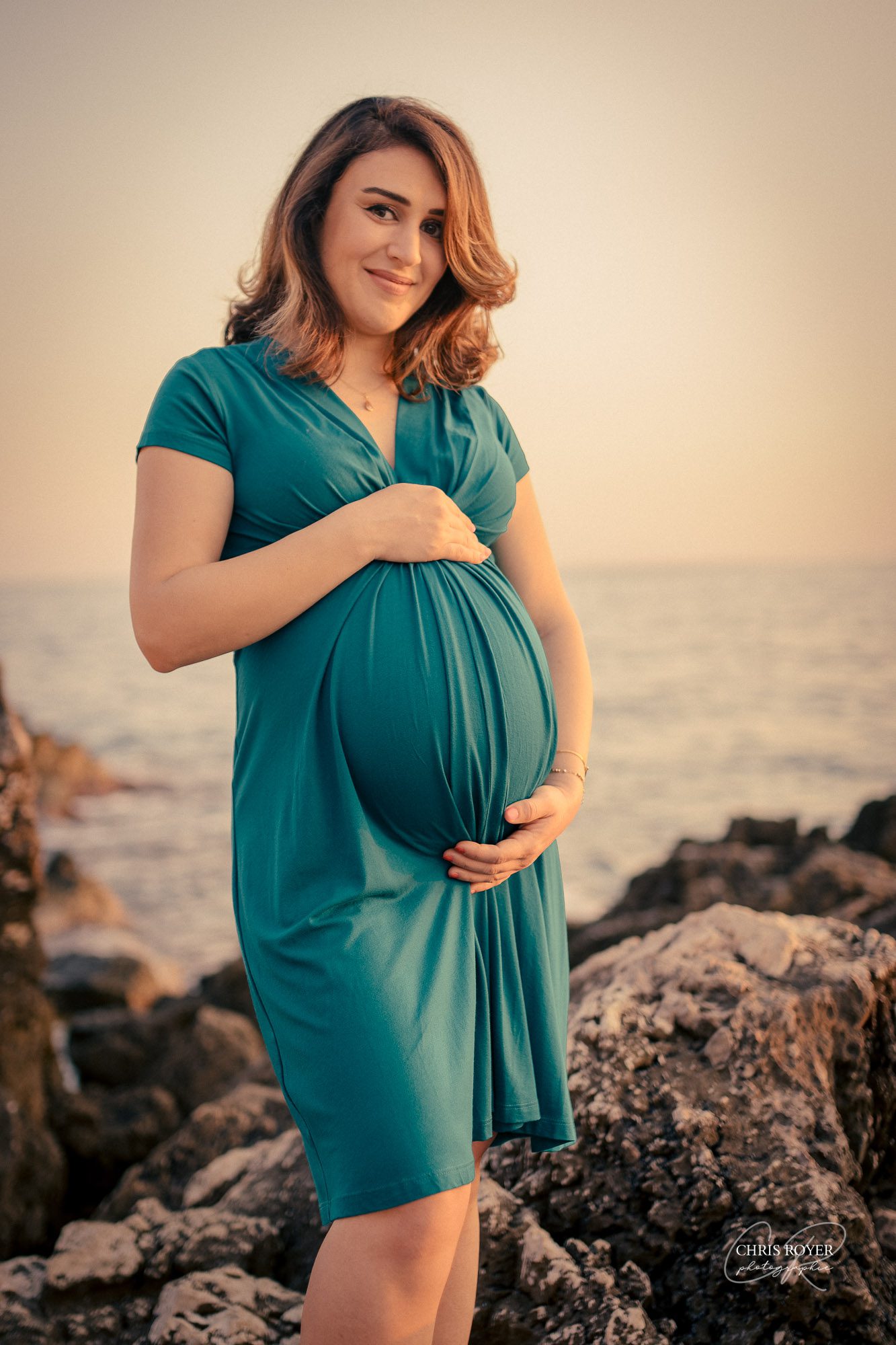 Une femme enceinte vêtue d'une robe sarcelle se tient sur un terrain rocheux au bord de la mer, au coucher du soleil dans les Alpes-Maritimes, caressant doucement son ventre et souriant à l'appareil photo, capturant ainsi un beau moment de maternité.