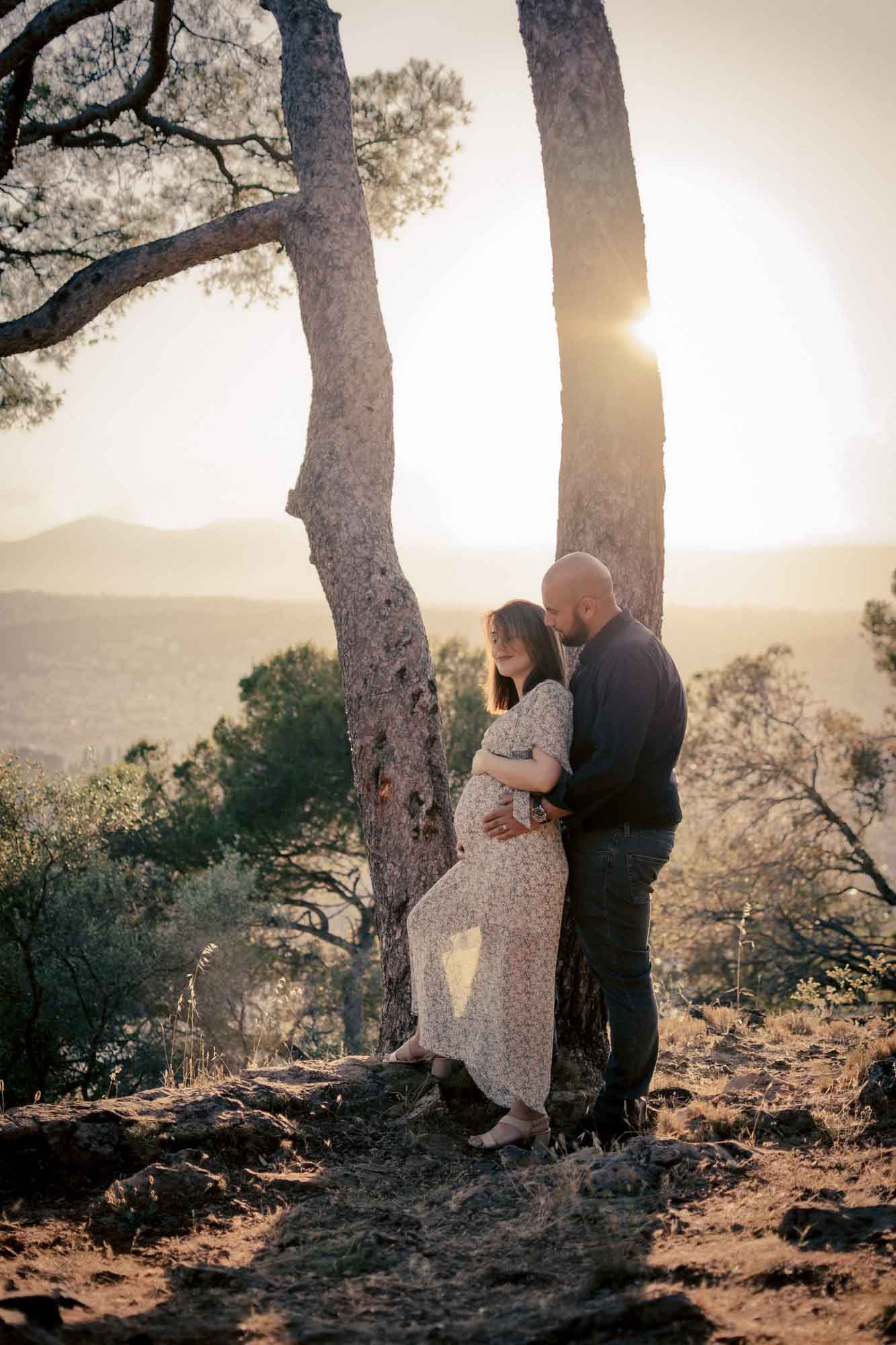 Couple attendant leur bébé enlacé entre deux pins, séance photographe grossesse Nice en hauteur au coucher du soleil.