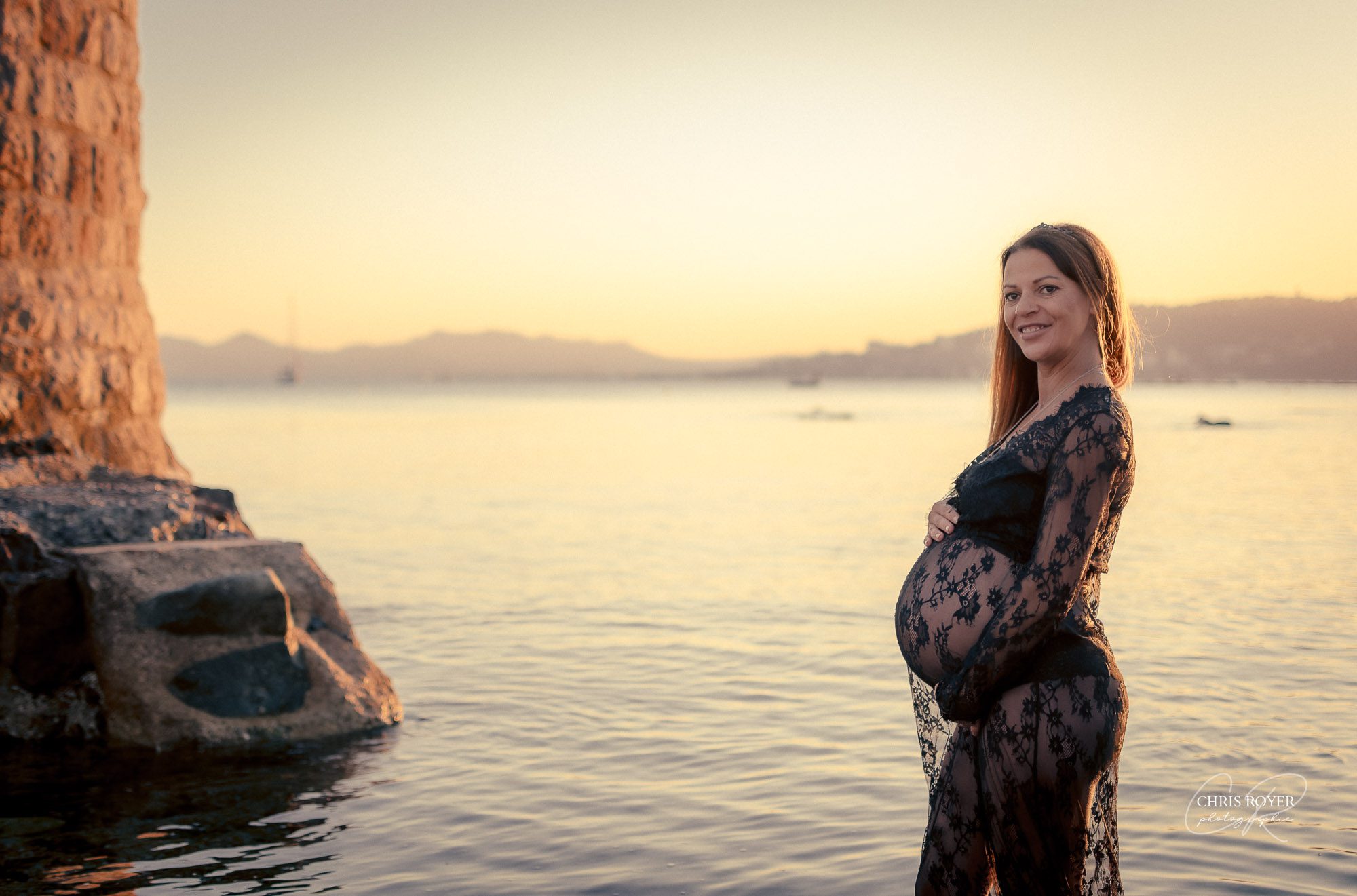 Une femme enceinte vêtue d'une robe en dentelle noire se tient dans une eau peu profonde près de gros rochers au coucher du soleil, souriant et tenant son ventre - un moment de maternité sereine parfait pour un portfolio de grossesse. L'eau calme et les collines lointaines scintillent sous le ciel doré.