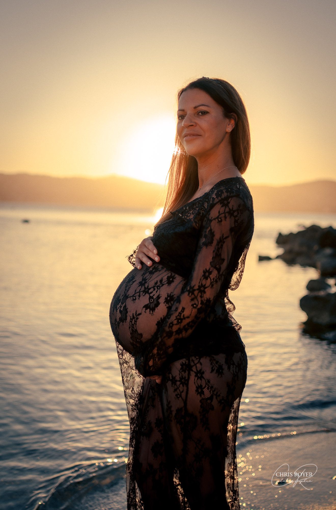 Une femme enceinte se tient au bord de l'eau au coucher du soleil à Nice, vêtue d'une robe en dentelle noire transparente et tenant doucement son ventre, avec les montagnes et les vagues tranquilles en arrière-plan - un ajout élégant à tout portefeuille de grossesse.