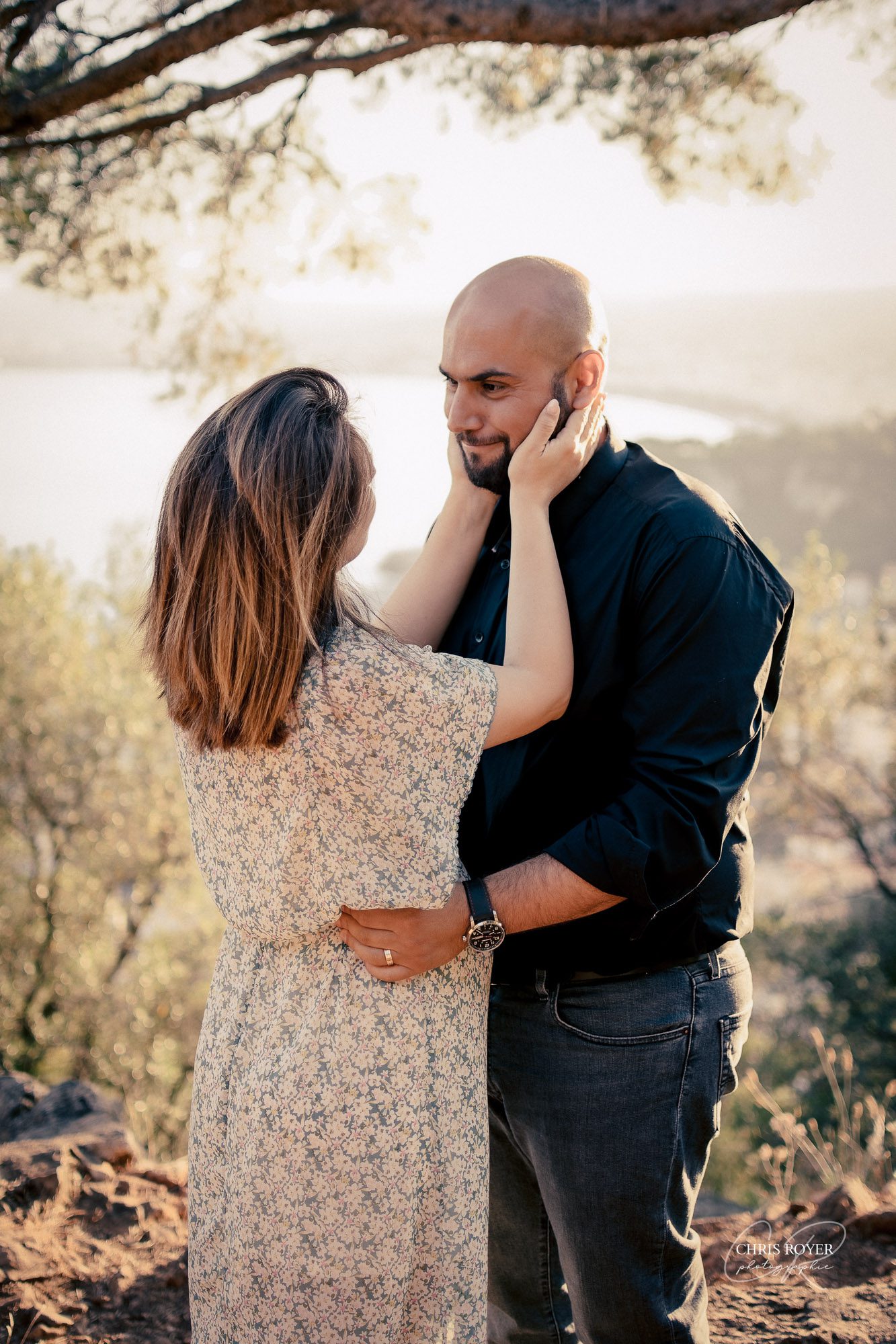 Un couple se tenant par la taille, échangeant un regard tendre en pleine nature, avec en arrière-plan la ville de Nice. Lors de leur séance de photo de grossesse.
