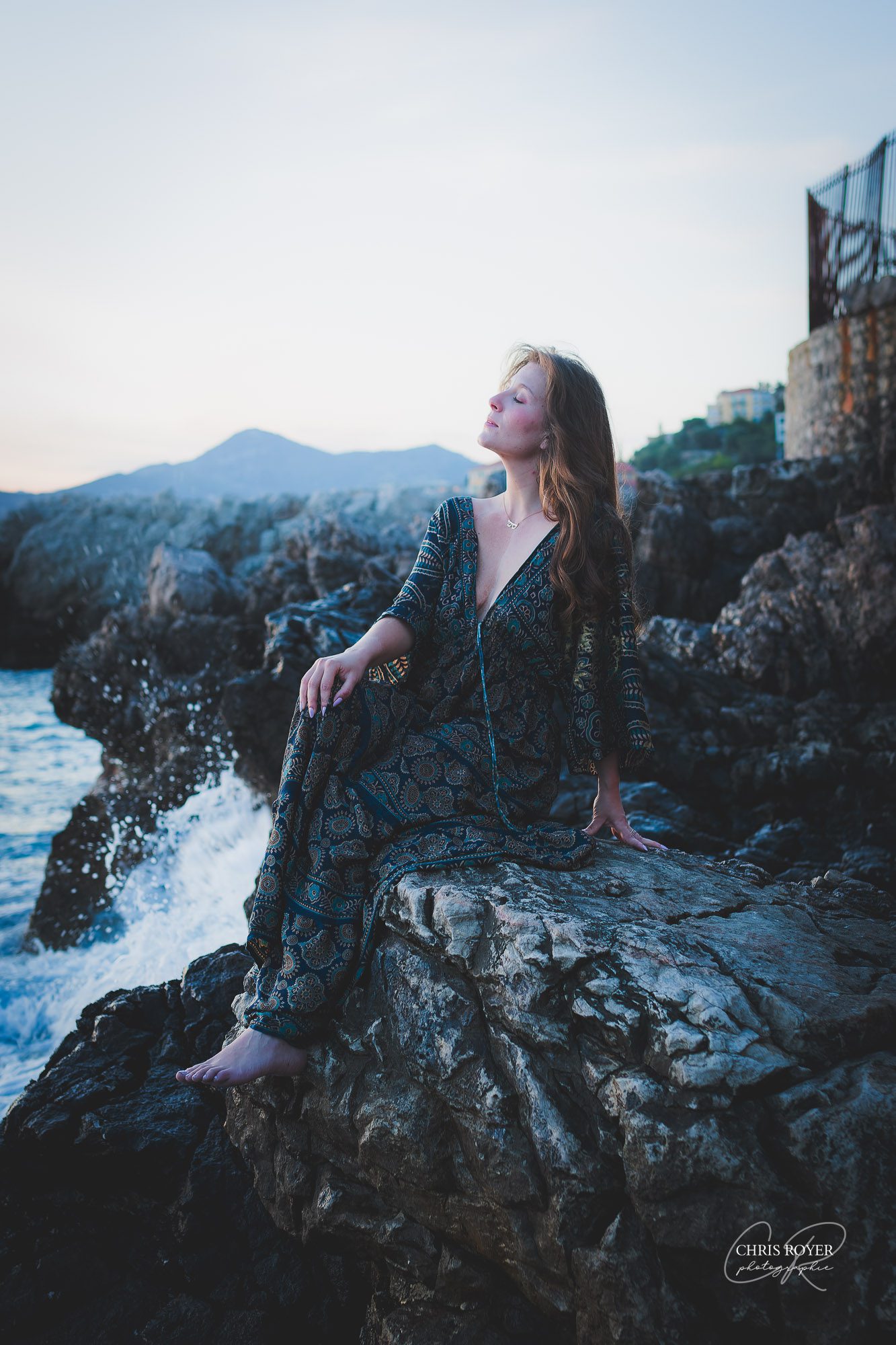 Une femme vêtue d'une robe à motifs est assise pieds nus sur des falaises rocheuses en bord de mer près de Nice, regardant vers le haut les yeux fermés tandis que les vagues de l'océan s'écrasent à proximité - parfait pour un portfolio de portraits naturels Nice, avec des montagnes et des bâtiments en arrière-plan sous un ciel bleu.