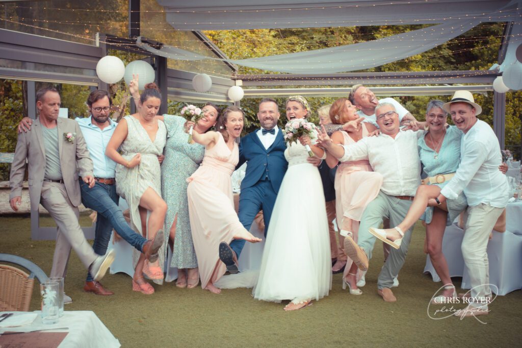 Une joyeuse fête de mariage, comprenant les mariés, pose ensemble en plein air pour une photo mariage émotions animée, en riant et en levant une jambe. Tout le monde est habillé en tenue de soirée, avec des couleurs claires et des expressions joyeuses.