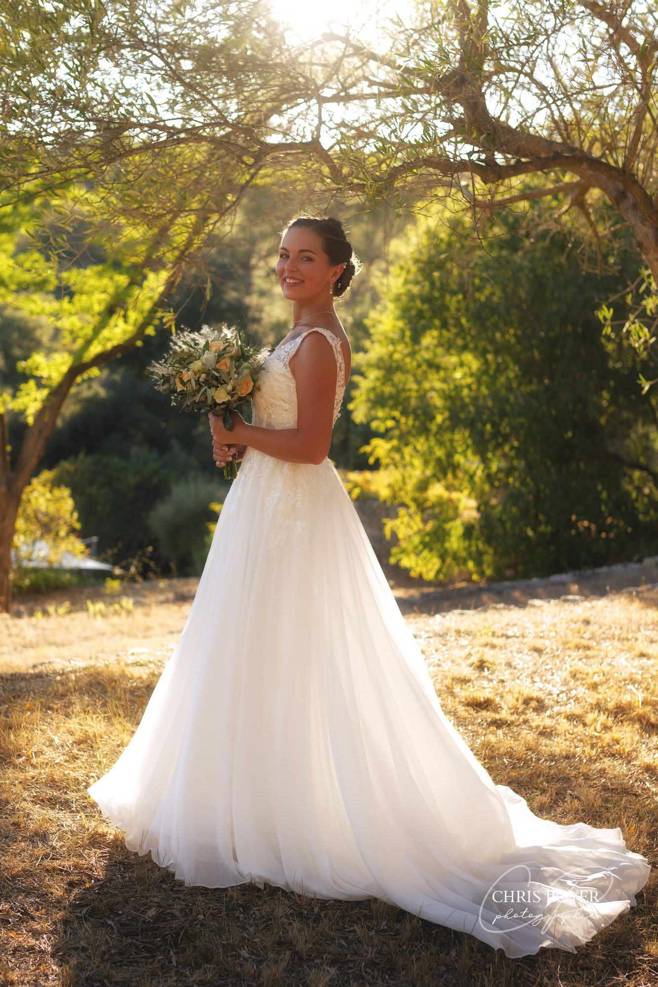 Mariée en robe blanche tenant son bouquet de fleurs dans un jardin arboré lors d’une séance photo de mariage à Grasse