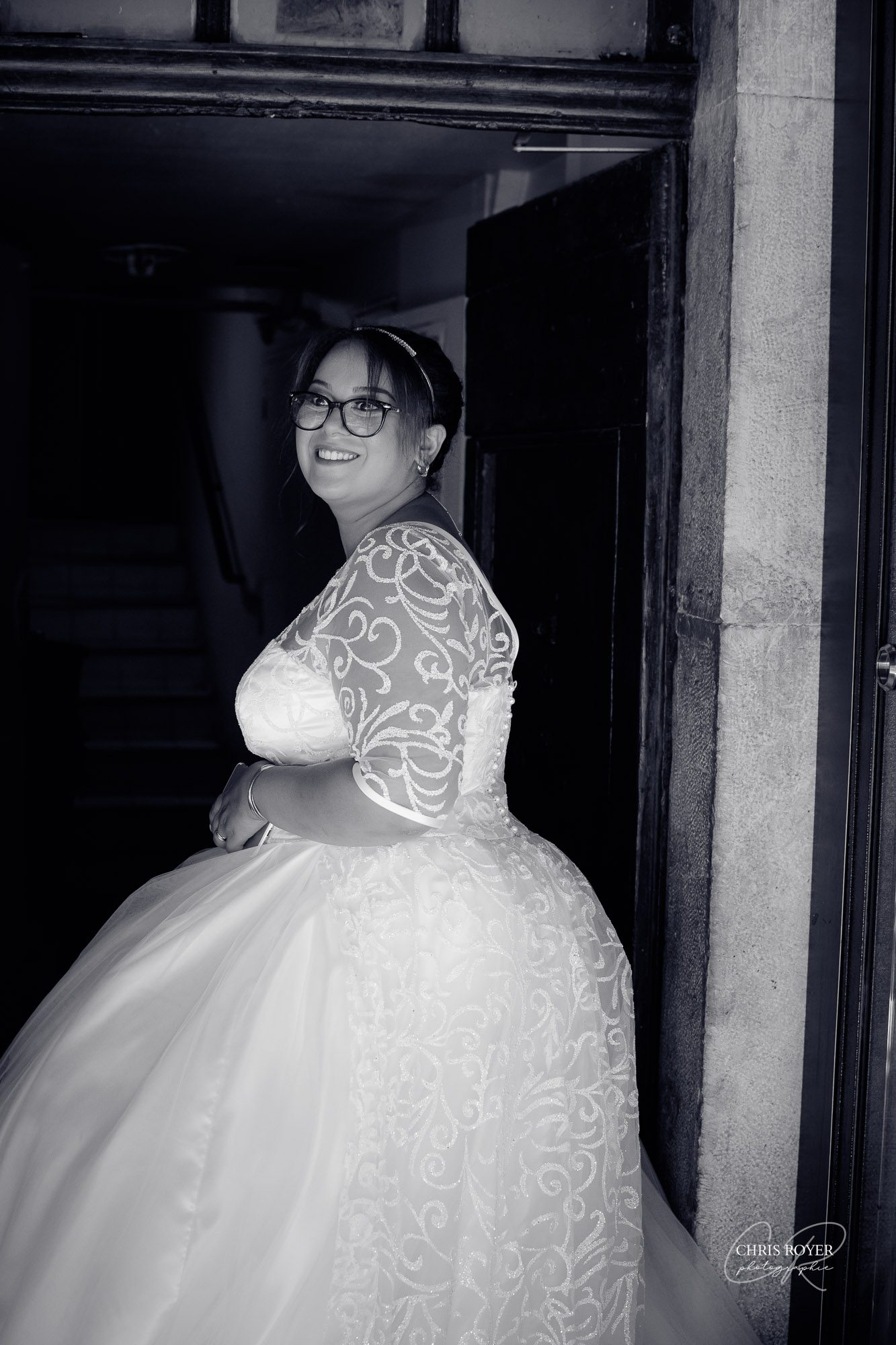 Une mariée en robe blanche avec des manches en dentelle et des lunettes se tient souriante dans l'embrasure d'une porte, en regardant par-dessus son épaule. Capturée en noir et blanc, cette photo évoque l'élégance intemporelle d'un reportage mariage Mougins.