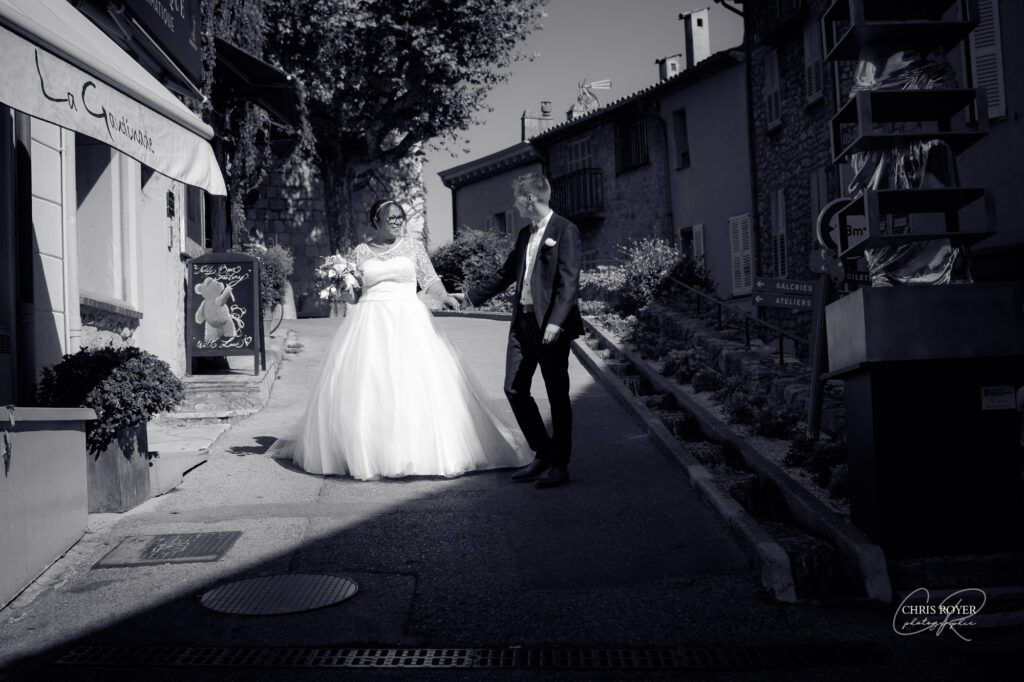 Une mariée en robe blanche et un marié en costume marchent ensemble dans une rue étroite et ensoleillée d'un village pittoresque, entourés de bâtiments en pierre et d'enseignes extérieures - parfait pour un reportage mariage Mougins aux couleurs chaudes.