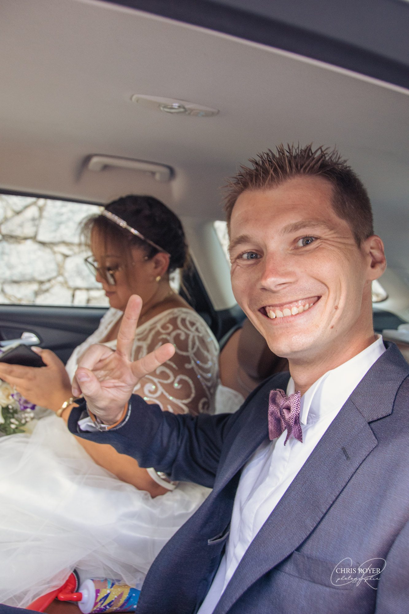 Un homme souriant en costume fait le signe de la paix, assis dans une voiture. À ses côtés, une mariée en robe de mariée et diadème consulte son téléphone. La lumière du soleil entre par la fenêtre, parfaitement capturée dans le cadre d'un reportage mariage Mougins.