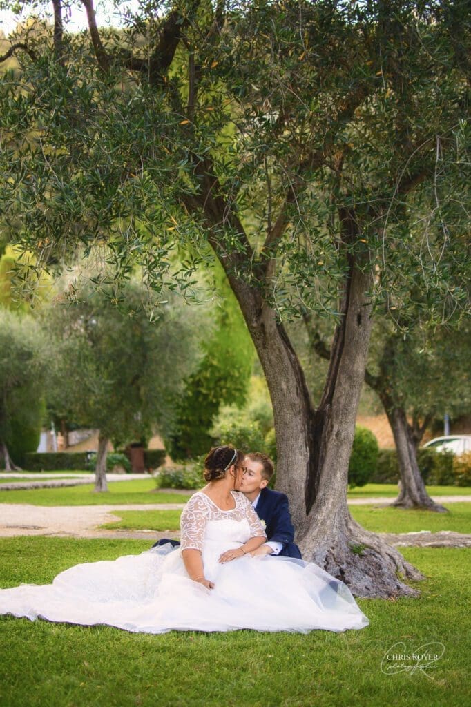 Une mariée et un marié sont assis sur l'herbe sous un grand arbre, le marié embrassant doucement la joue de la mariée. Entourés d'une végétation luxuriante, ce moment capture la romance d'un reportage mariage Mougins par un photographe mariage Mougins.
