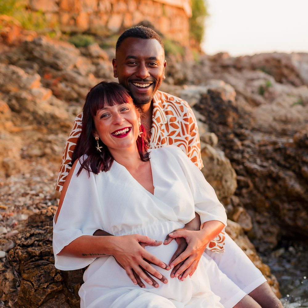 Un couple souriant est assis sur des rochers en plein air, la femme en robe blanche tenant son ventre de femme enceinte et l'homme derrière elle. Tous deux capturé par une photographe de Nice.