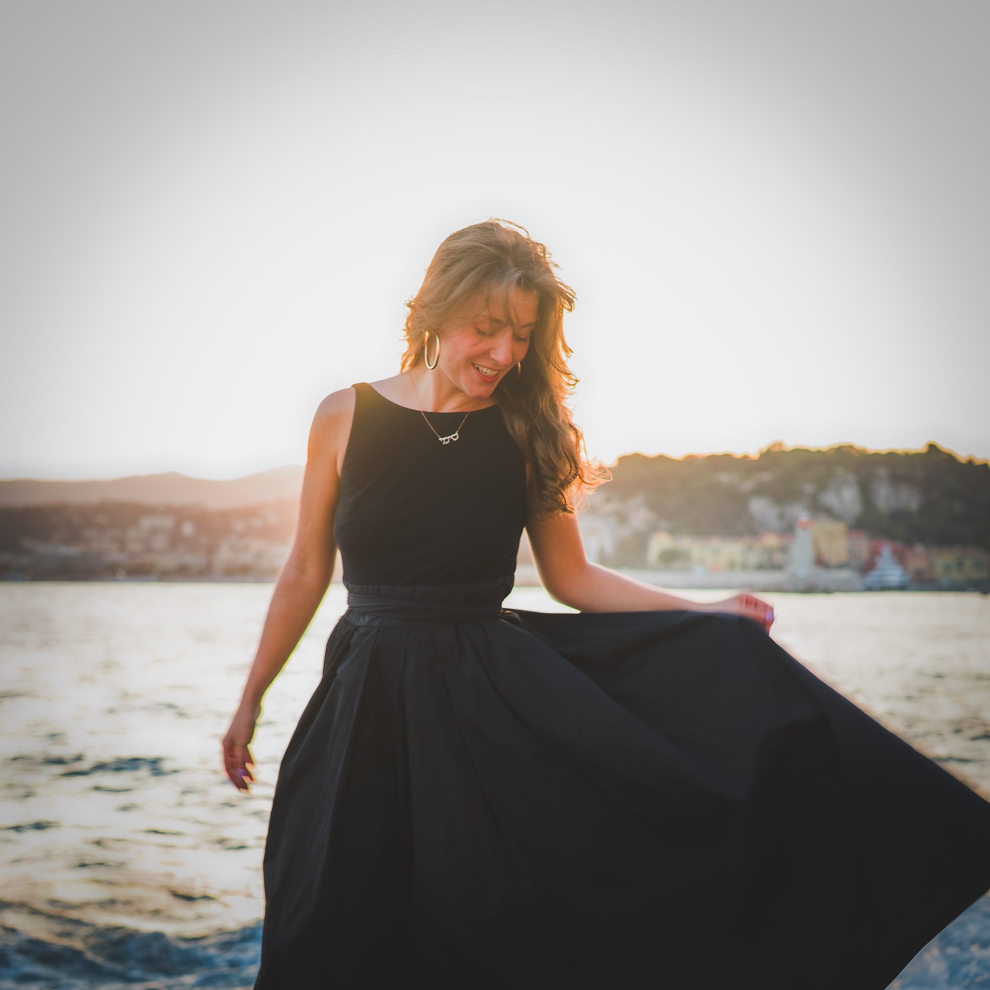 Une femme en robe noire se tient au bord de l'eau au coucher du soleil, souriant en tenant sa jupe pendant une séance photo portrait lifestyle naturel à Nice.