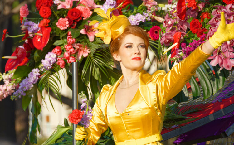 Une femme vêtue d'une robe et d'un chapeau jaune vif se tient devant une grande exposition florale au Carnaval de Nice, tenant un bâton et tendant une main gantée au milieu de fleurs roses et rouges.