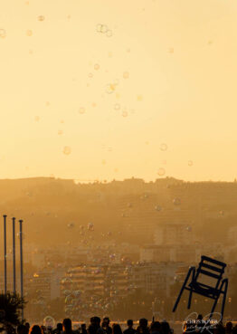 Silhouette d'une foule avec des bulles flottantes au coucher du soleil et une grande sculpture de chaise de plage de Nice en arrière-plan.
