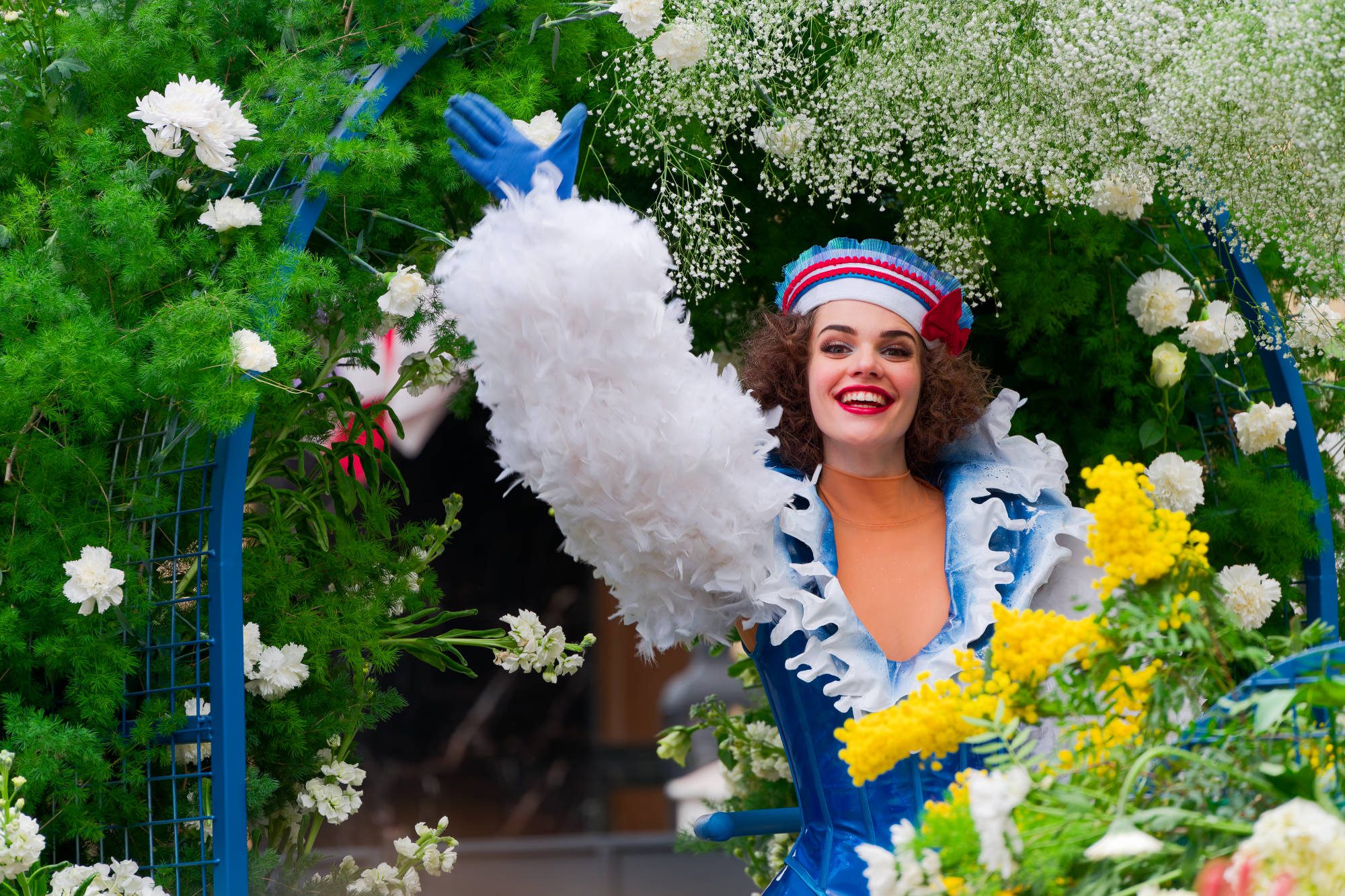 Une femme vêtue d'un costume coloré avec des manches en plumes blanches et un chapeau de marin sourit et salue parmi les fleurs et la verdure, capturant l'esprit festif du Carnaval de Nice Parcours photographique Nice.