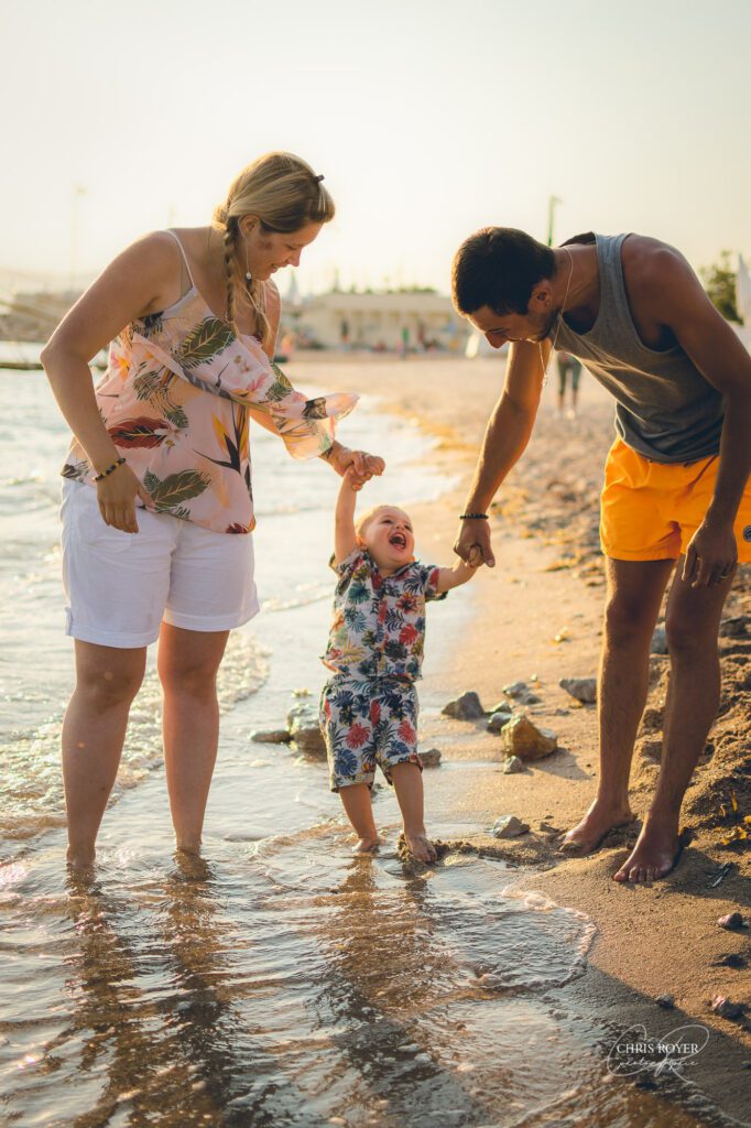 Séance photo famille lifestyle plage Palm Beach Cannes. Des parents tenant la main de leur fils au bord de la mer au coucher du soleil, moment de complicité tendre pour un bonheur familial authentique !