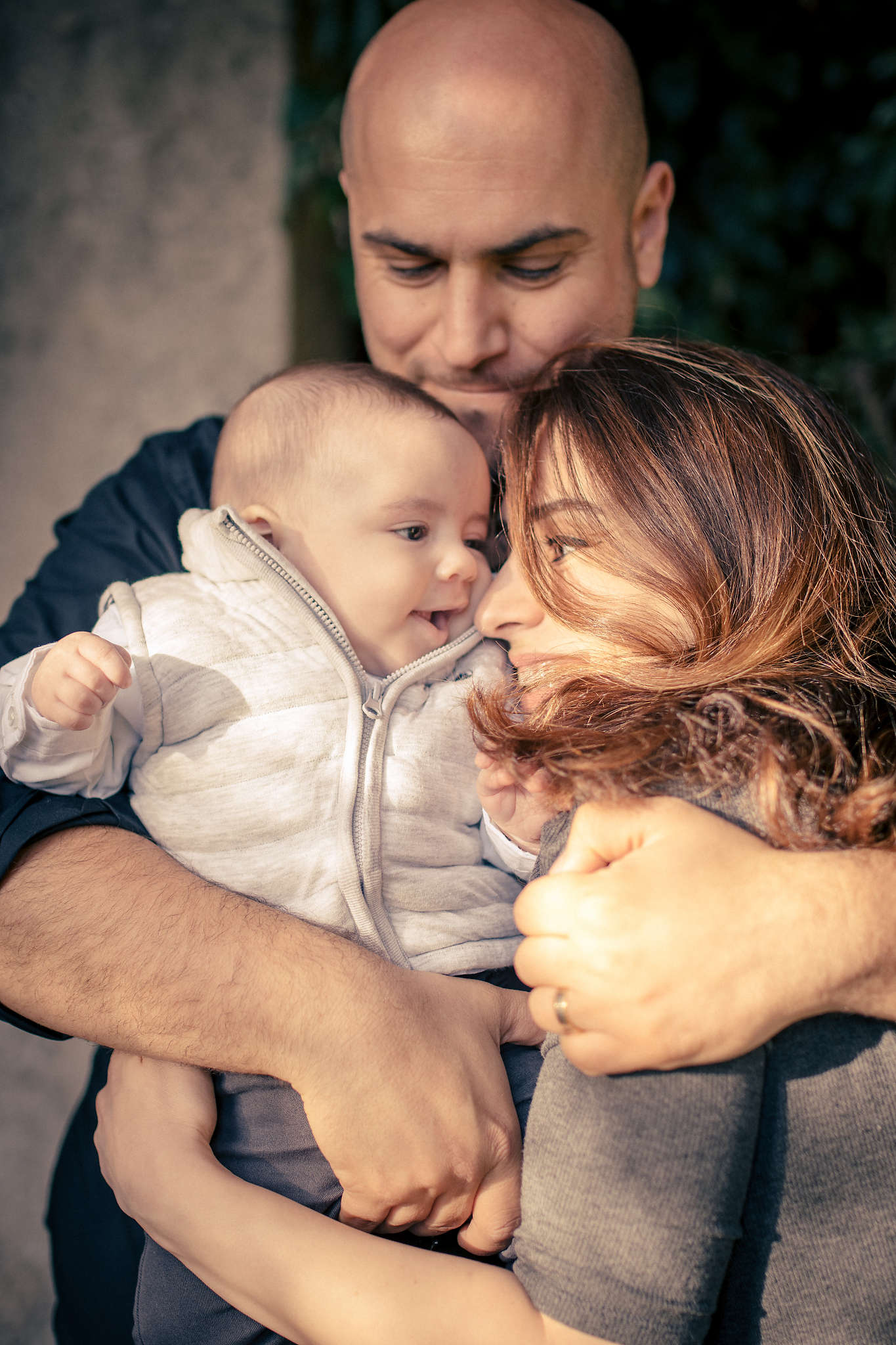 Un homme et une femme s'embrassent en tenant un bébé, tout en souriant en regardant leur enfant dans une tenue de couleur claire. Un moment réconfortant capturé par un photographe lifestyle Nice.