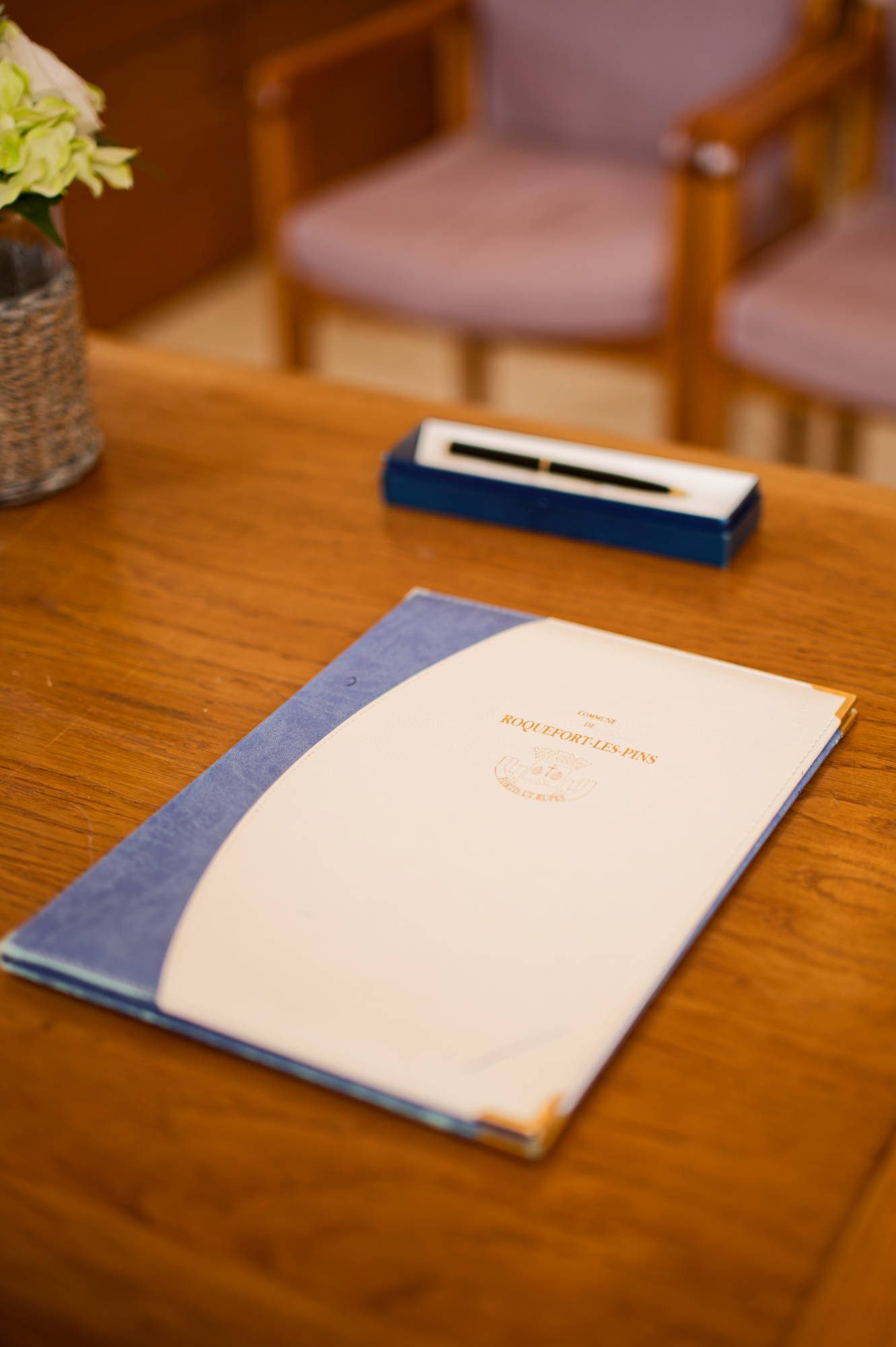 Un dossier fermé étiqueté Réceptionniste repose sur un bureau en bois, à côté d'un document porte mariage roquefort-les-pins, avec un stylo dans un support et deux chaises à l'arrière-plan.
