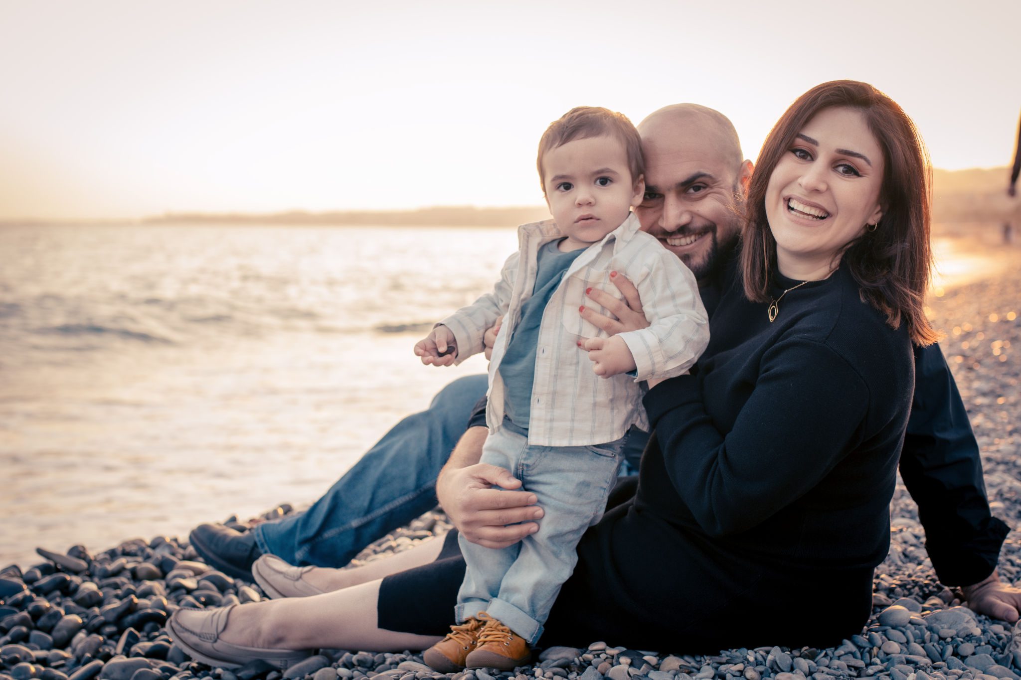 Une famille de trois personnes est assise sur la promenade des anglais au coucher du soleil. L'enfant est assis sur les genoux du père et la mère sourit à l'appareil photo.
