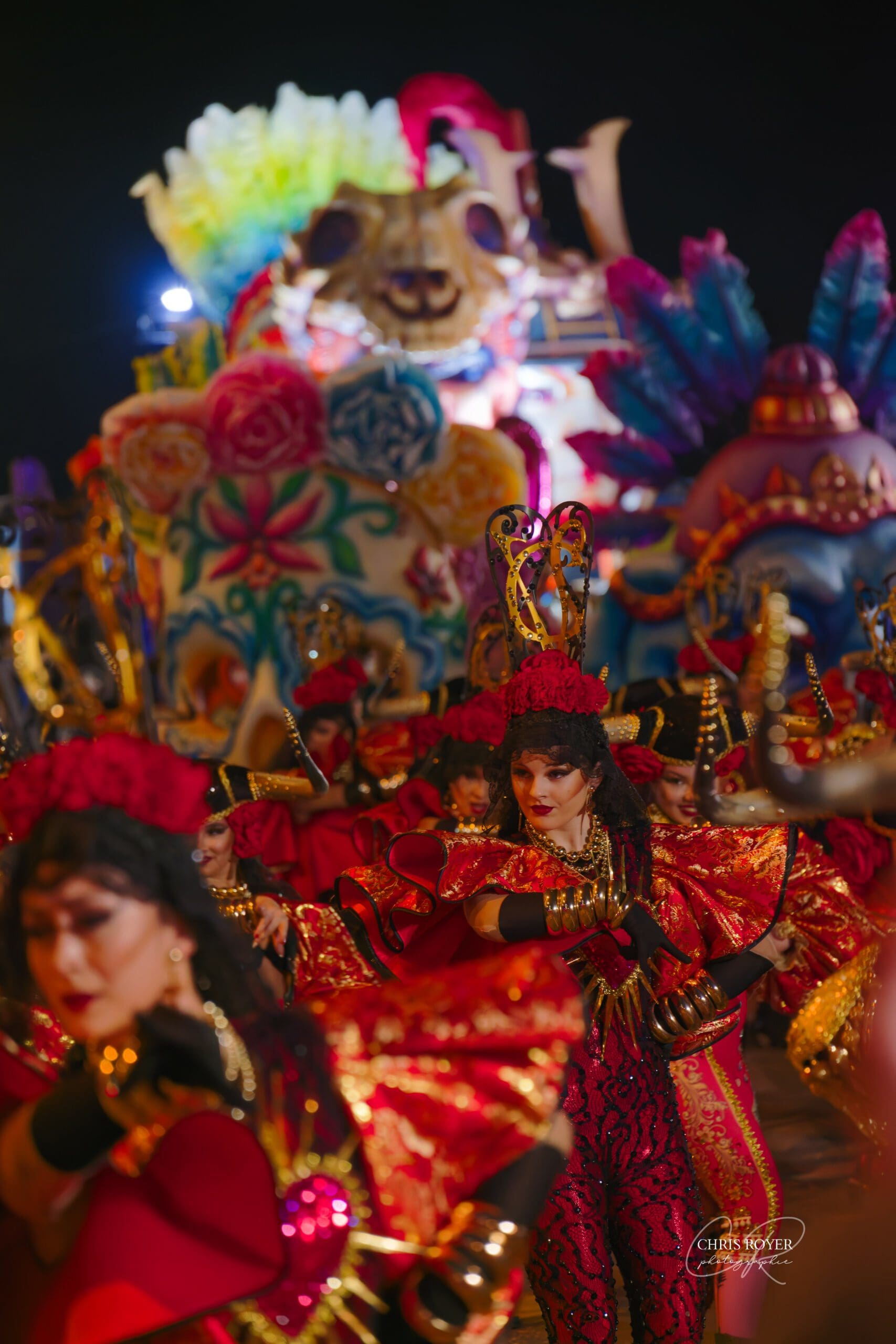 Danseuses en costume rouge et or défilant de nuit devant un char multicolore et illuminé au Carnaval de Nice 2026.