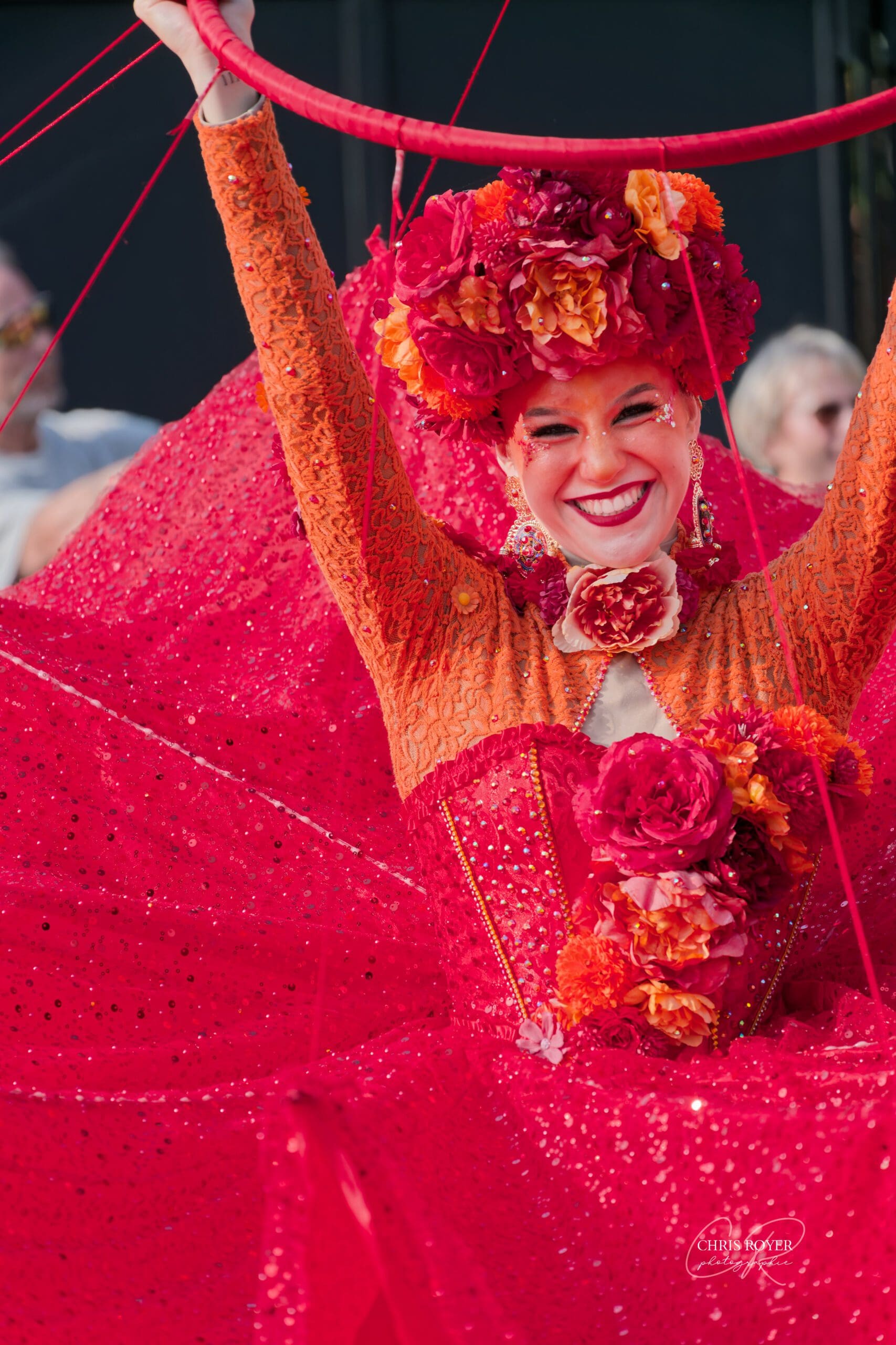 Artiste du Carnaval de Nice 2026 souriante, en costume rouge orangé pailleté brandissant un cerceau au-dessus de sa tête.