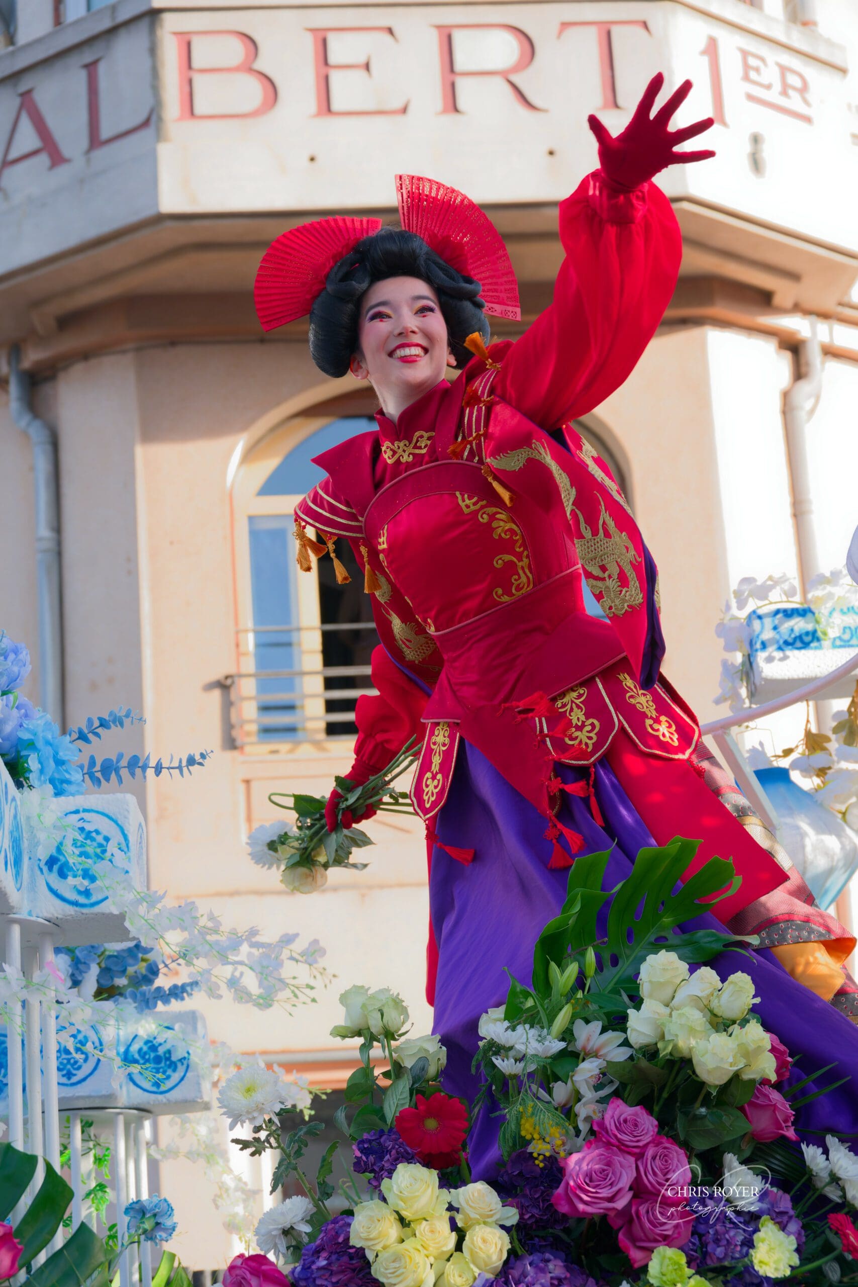 Artiste du Carnaval de Nice 2026 en costume écarlate luxueux avec coiffe ornementale de thème Chinois, souriant face, bras levé après avoir lancé une rose sur un char décoré de fleurs colorées et près du Jardien Albert 1er de Nice.