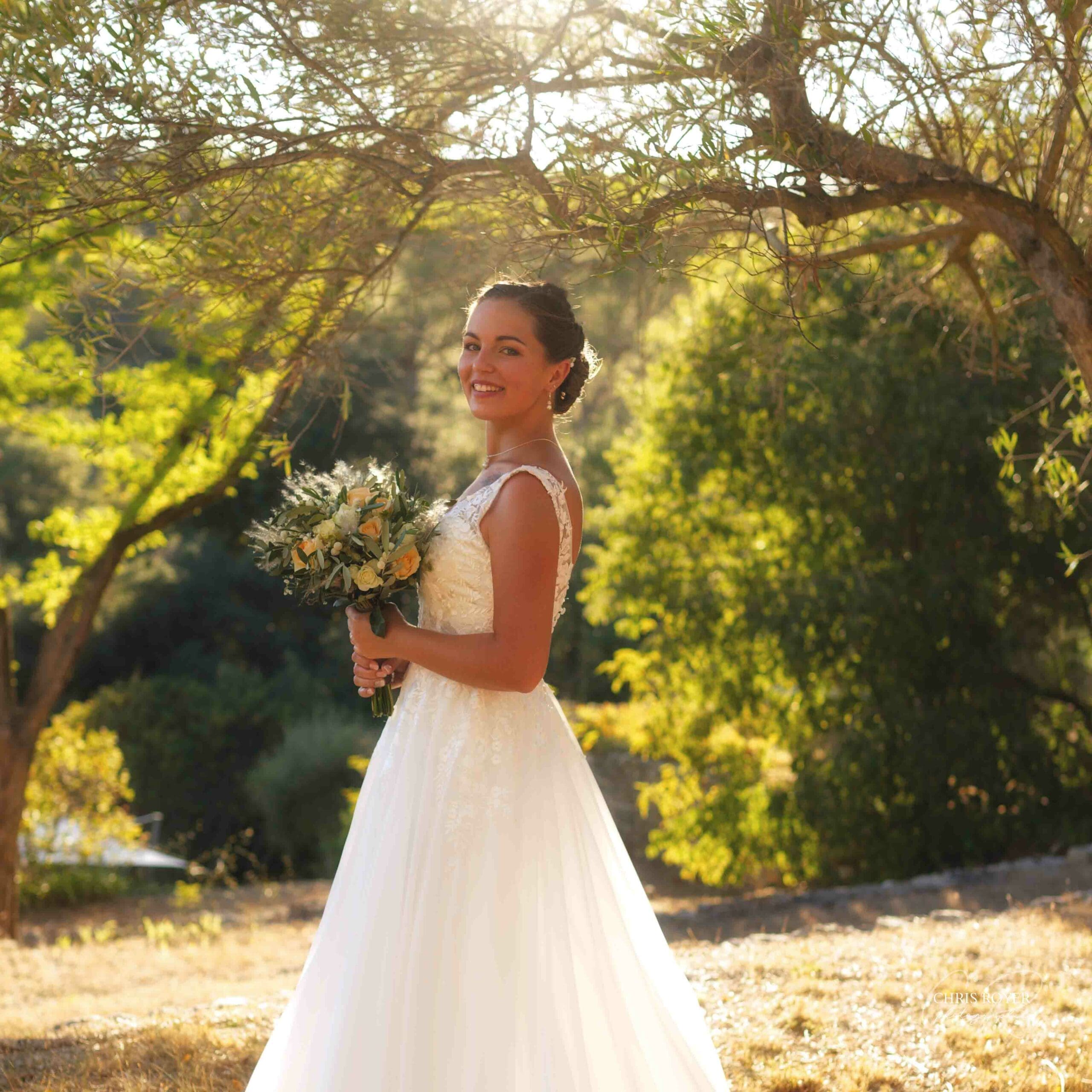 Mariée en robe de dentelle tenant un bouquet, photographiée en extérieur sous les arbres baignés de soleil, lors de son mariage.