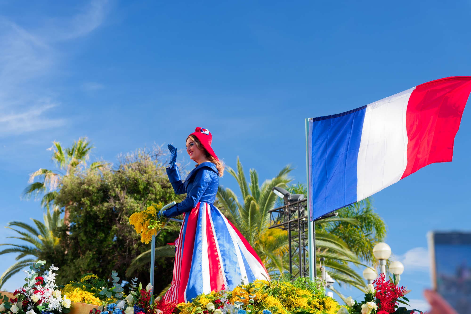 Ambassadrice en costume tricolore saluant la foule sur un char fleuri de mimosas lors du Carnaval de Nice 2026 sous le drapeau français
