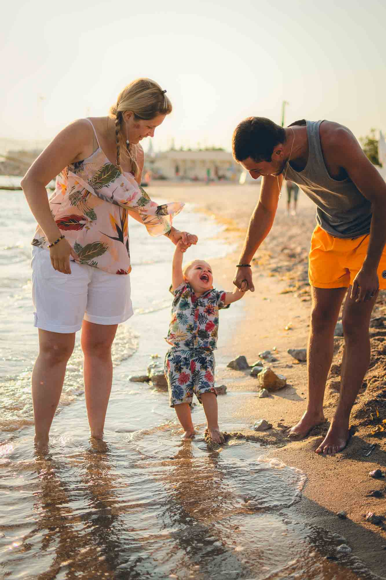 Parents et leur bébé qui rit les pieds dans l’eau sur la plage de Cannes, séance avec un photographe famille Cannes