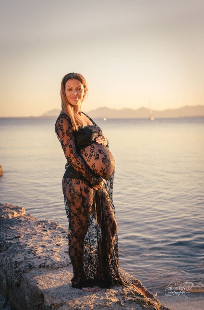 Une femme enceinte en robe noire en dentelle se tient sur une plage rocheuse au coucher du soleil, caressant tendrement son ventre avec ses deux mains. Cette image illustre une séance photo maternité réalisée par un photographe grossesse à Antibes, capturant l’intimité d’un moment précieux en extérieur.