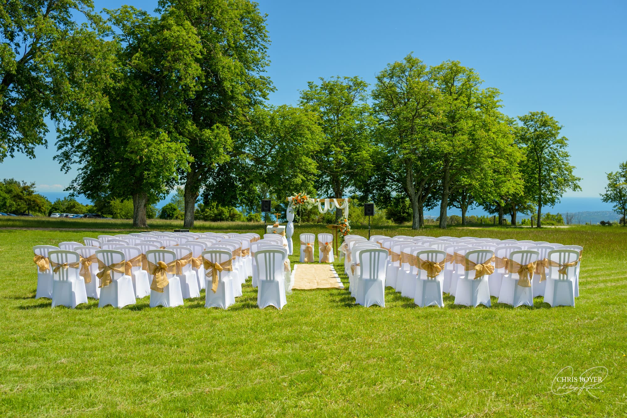 Cérémonie de mariage en extérieur au Domaine des Courmettes, avec une allée centrale bordée de chaises blanches ornées de rubans dorés, menant vers une arche florale sous un ciel bleu.