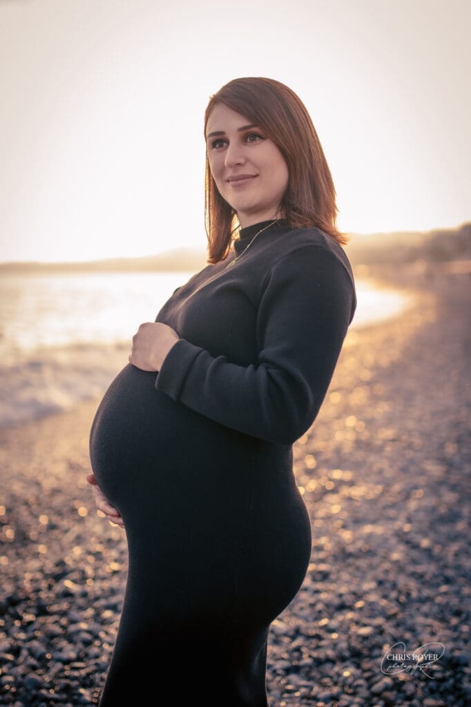 Femme enceinte debout sur une plage de galets au coucher du soleil, les mains posées sur son ventre arrondi, portant une robe noire par photographe grossesse à Nice: Chris Royer Photographie