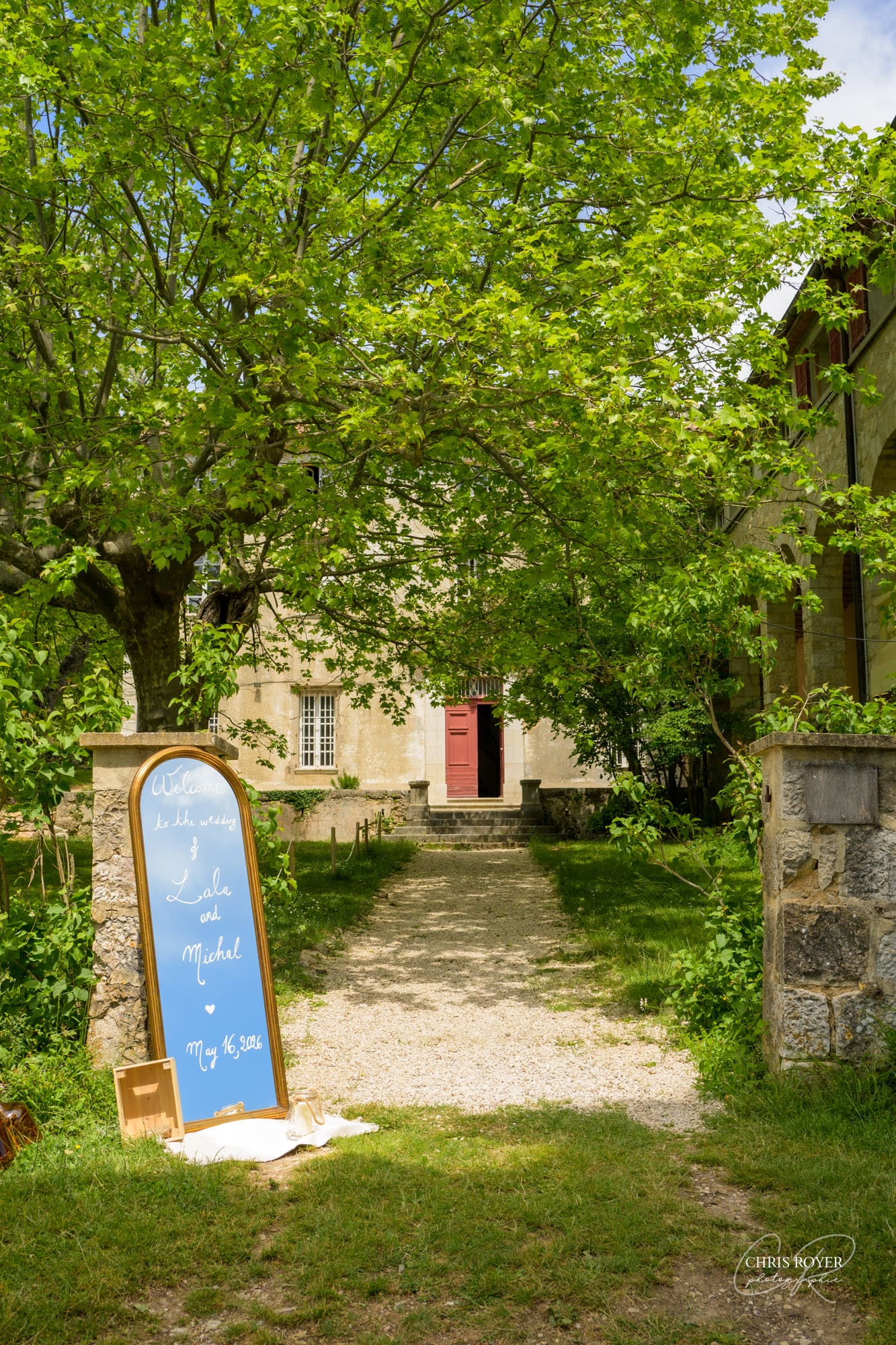 Panneau d'accueil de mariage pour Lala et Michal devant l'entrée en pierre du Domaine des Courmettes, porte rouge, chemin en gravier ombragé par un arbre.