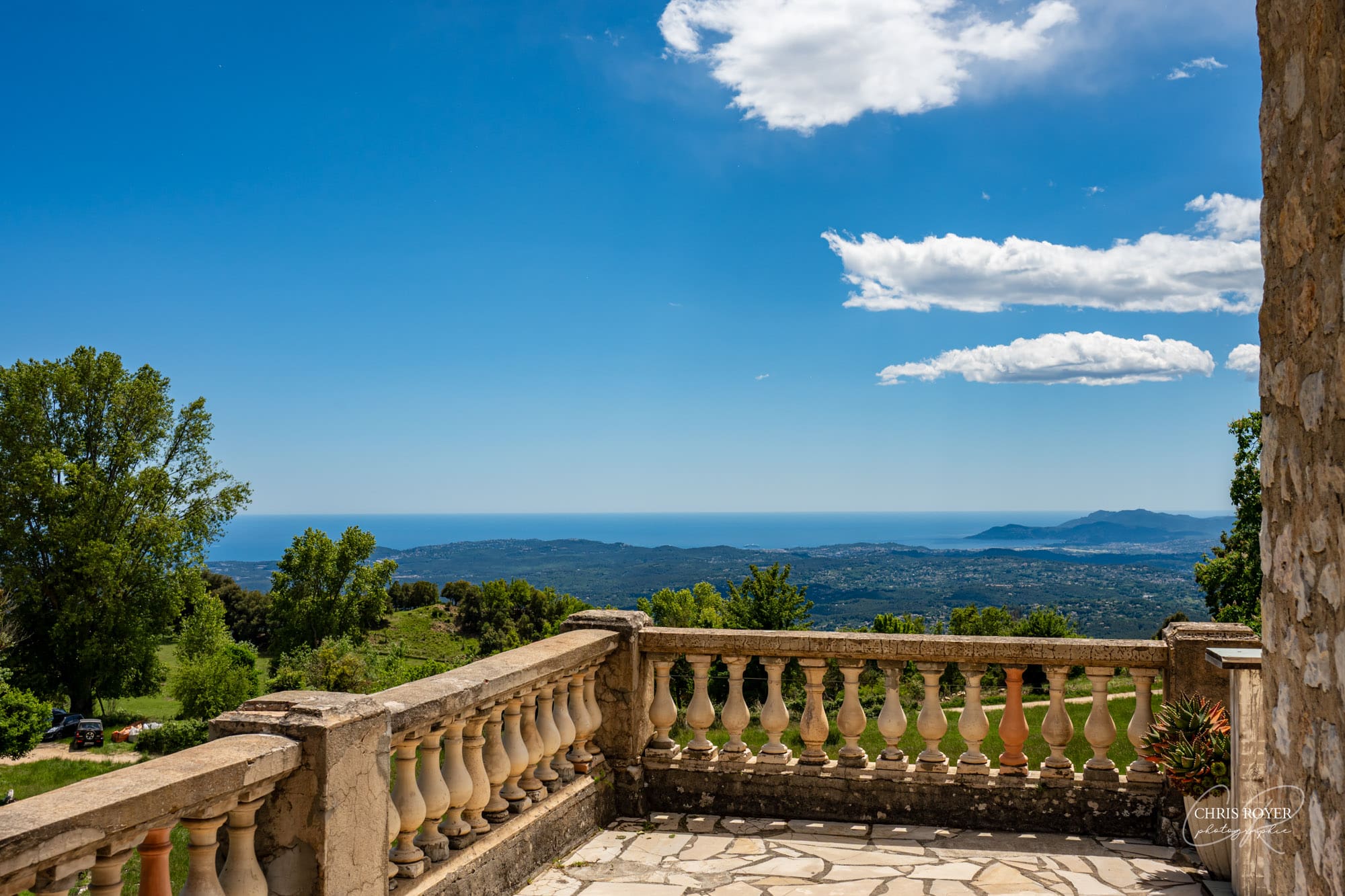 Vue panoramique depuis la terrasse en pierre du Domaine des Courmettes, balustrade à balustres classiques, ciel bleu avec nuages, mer Méditerranée et collines en arrière-plan.