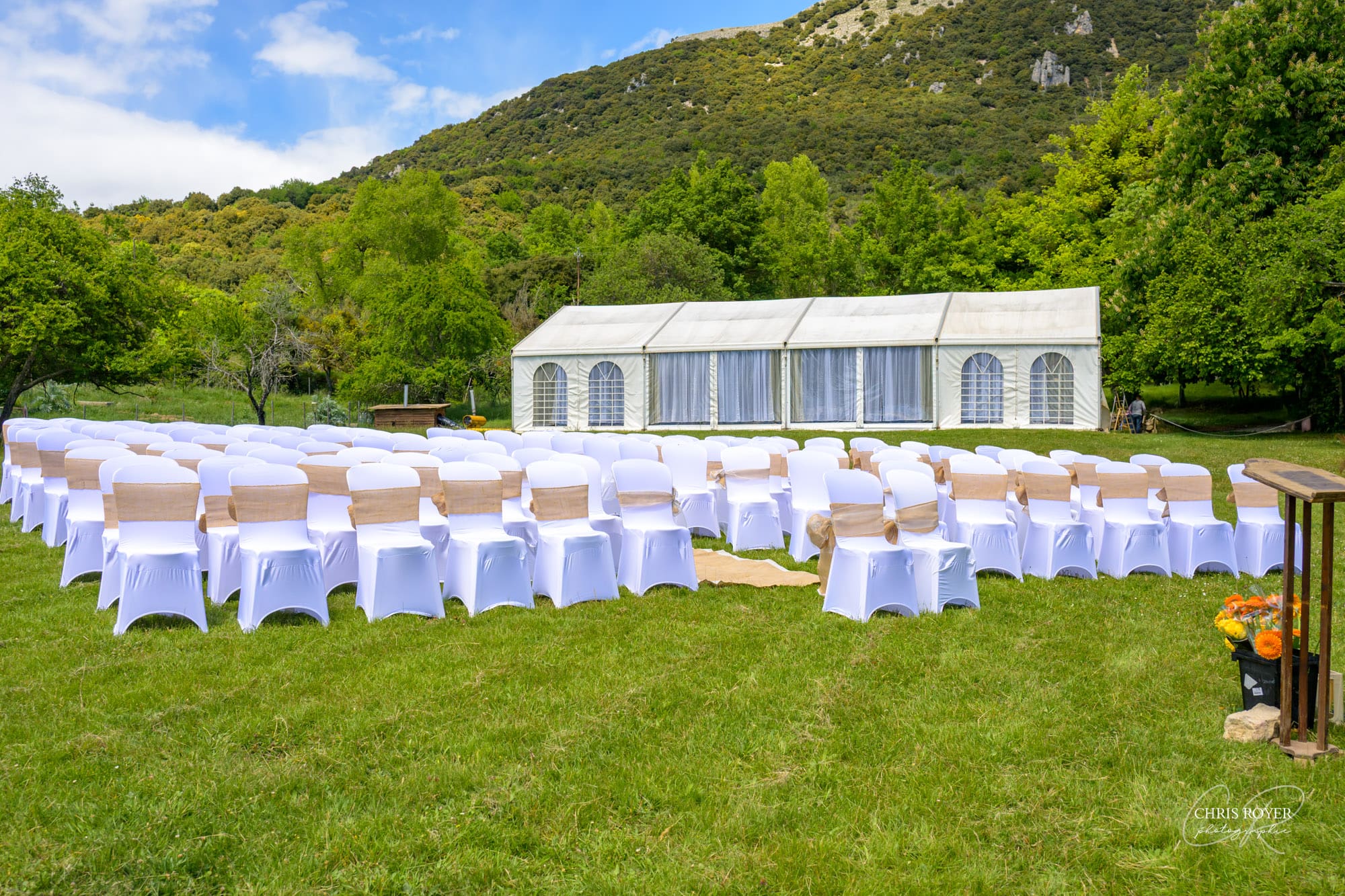 Vue d'ensemble du site de mariage au Domaine des Courmettes, rangées de chaises blanches sur une pelouse face à une tente de réception, avec les collines verdoyantes des Alpes-Maritimes en arrière-plan.