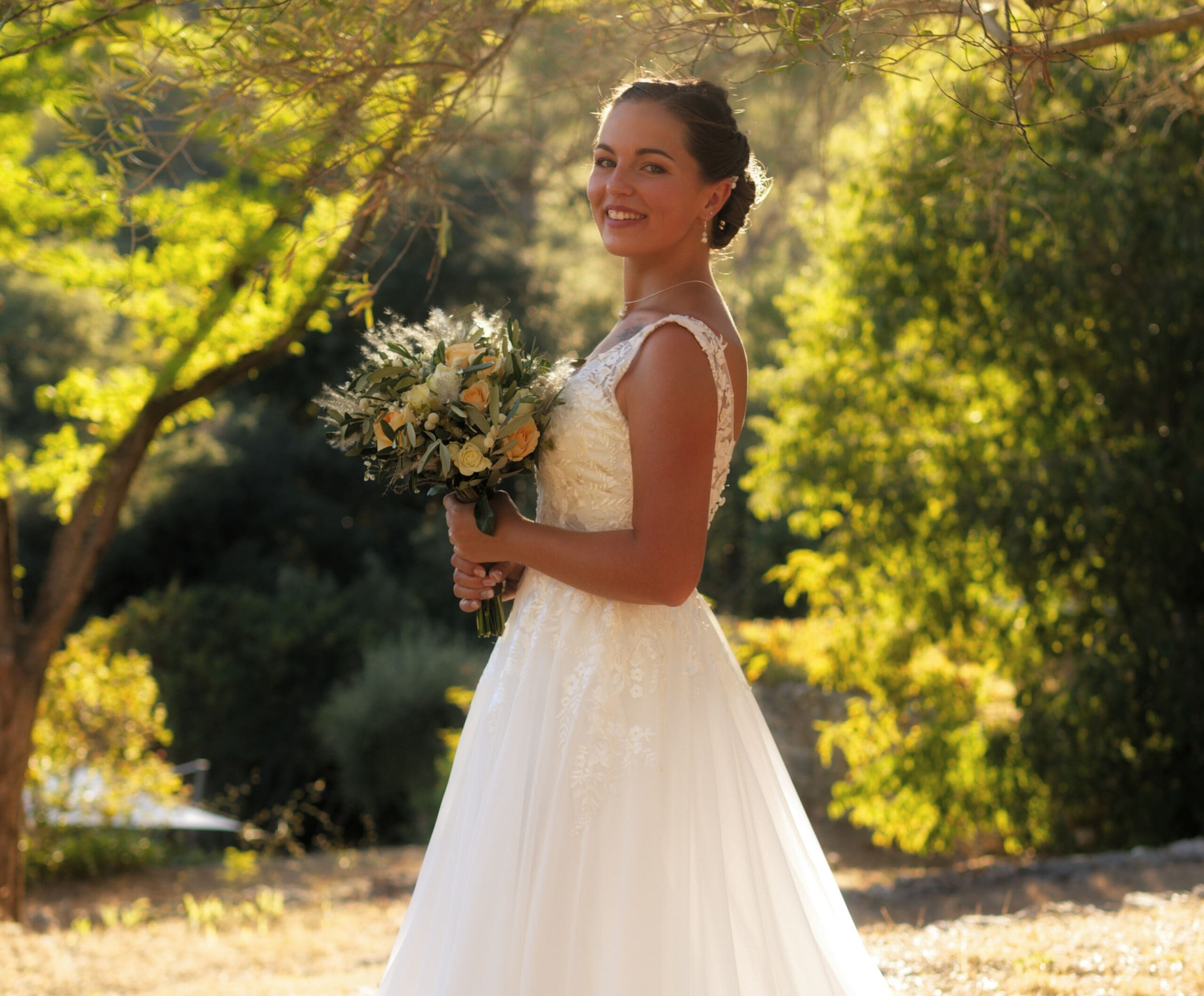 Mariée en robe blanche tenant son bouquet de fleurs dans un jardin arboré lors d’une séance photo de mariage à Grasse au Domaine de Tabourg