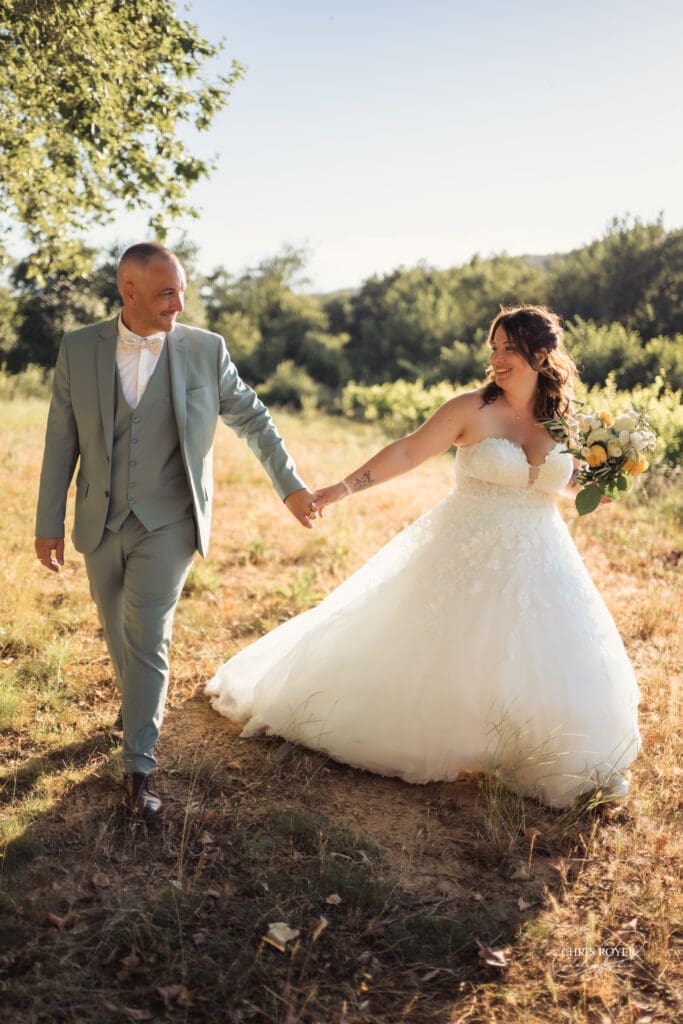 Mariés se tenant la main en marchant dans un champ ensoleillé, la mariée en robe de dentelle blanche tenant son bouquet. Mariés se tenant la main en marchant dans un champ ensoleillé, la mariée en robe de dentelle blanche tenant son bouquet.